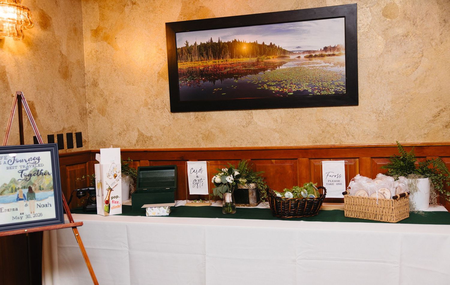Elegant wedding reception table with floral arrangements, gift baskets, and a framed landscape painting on a textured wall.
