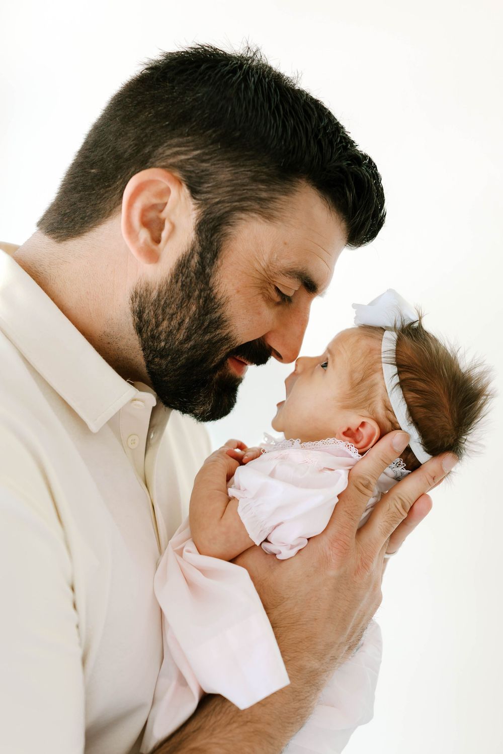 Bearded father tenderly touches noses with his newborn baby girl in a white bow headband.