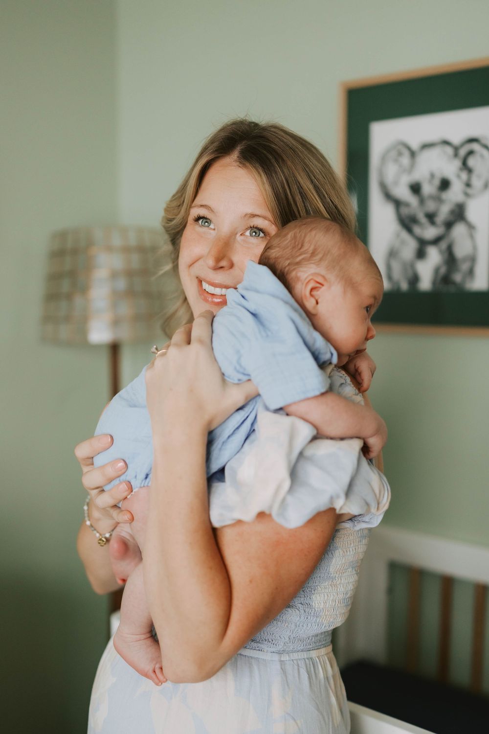 Smiling new mother in a light blue dress holds her sleeping newborn baby against her shoulder.