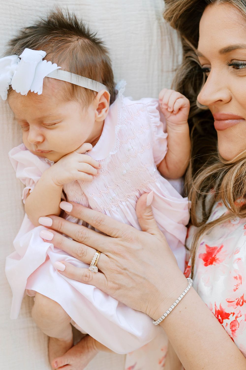 Mother in floral dress gazes lovingly at sleeping newborn girl in pink outfit with white bow.