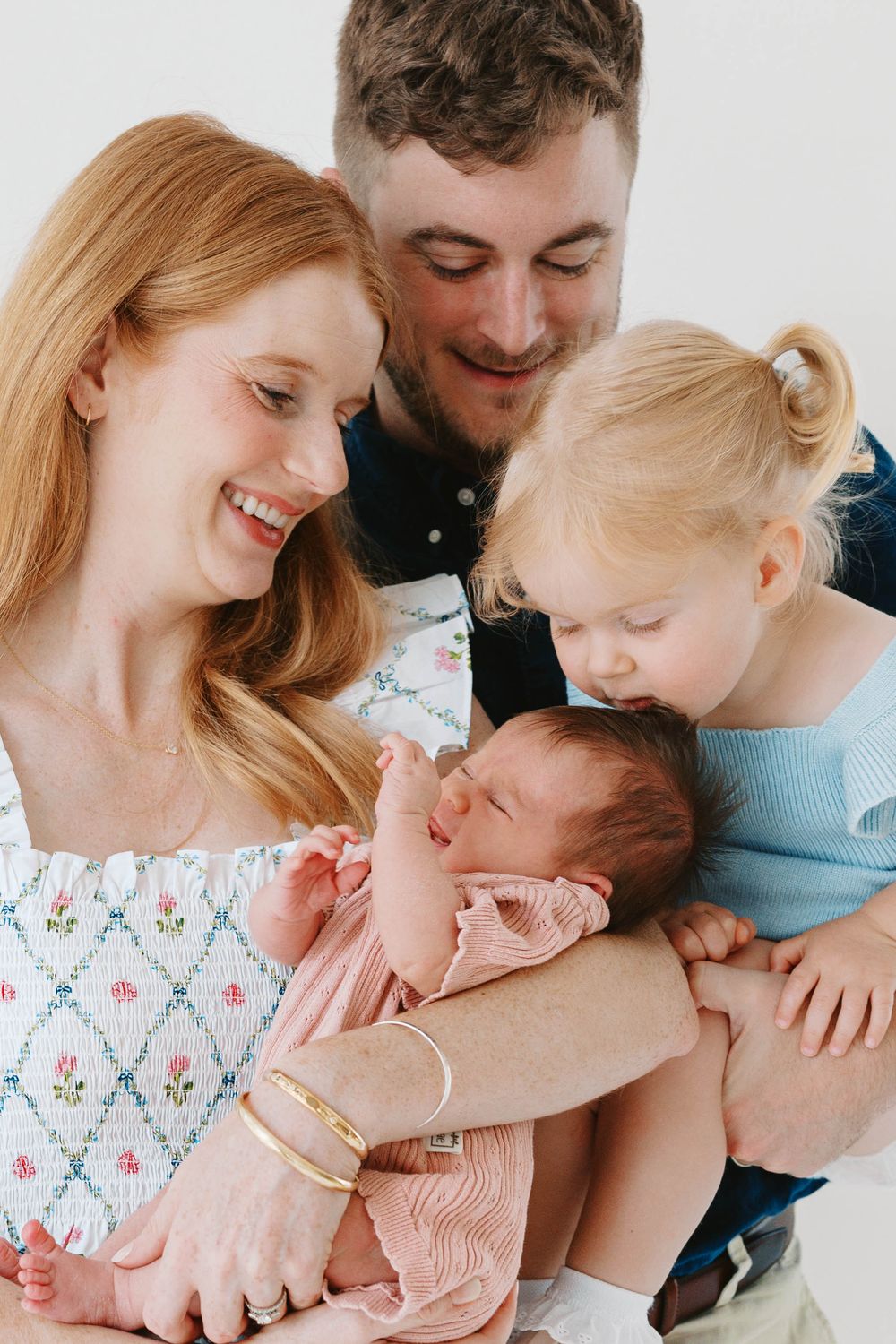 Happy family of four with redhead mother, father, toddler girl, and newborn baby in bright studio.