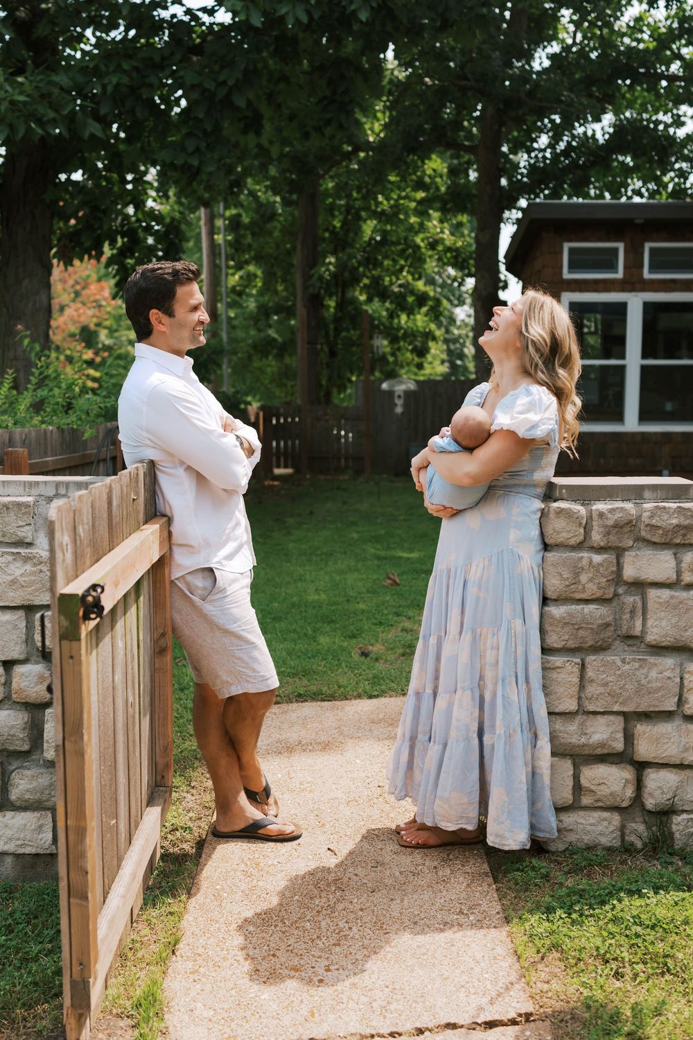 New parents stand at a garden gate, mother in blue dress holding newborn, father in white shorts.