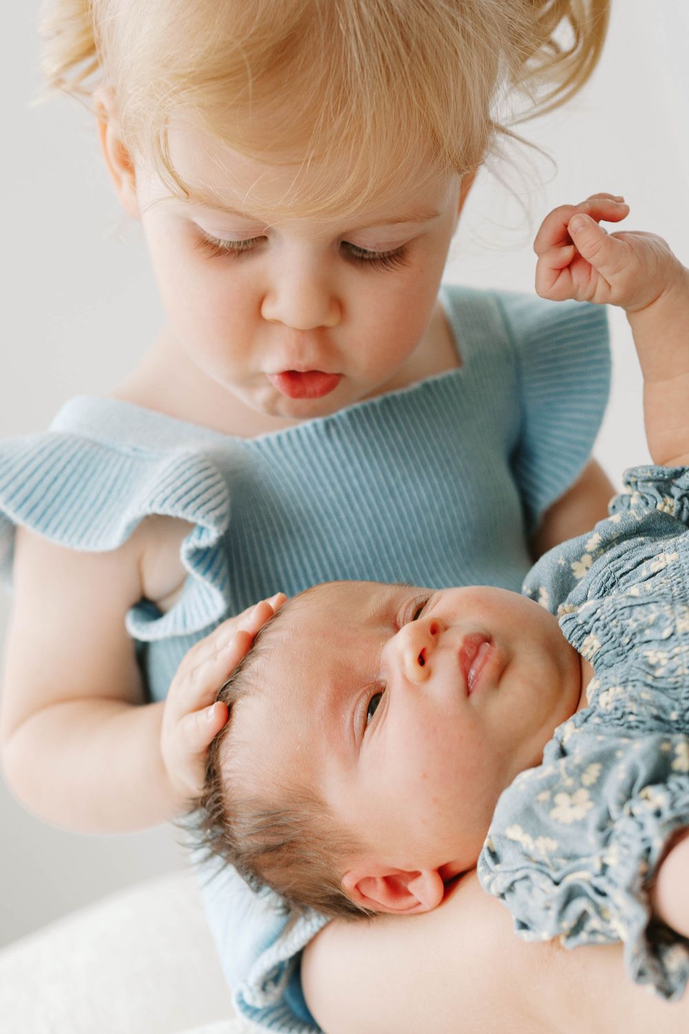 Curious blonde toddler girl in blue dress gently examines and holds a sleeping newborn baby.