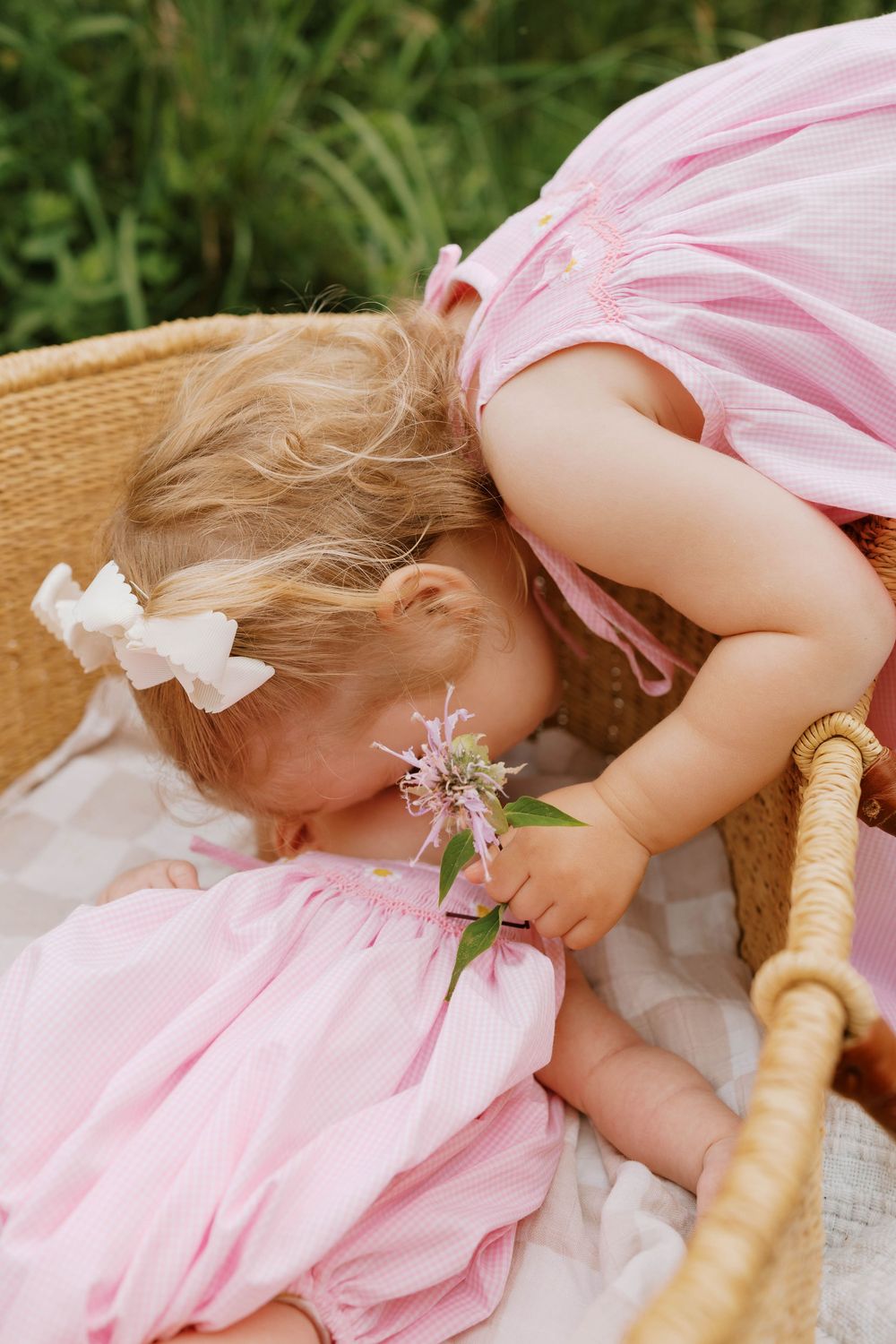 Two toddler girls in matching pink dresses lean together over a wicker basket outdoors.