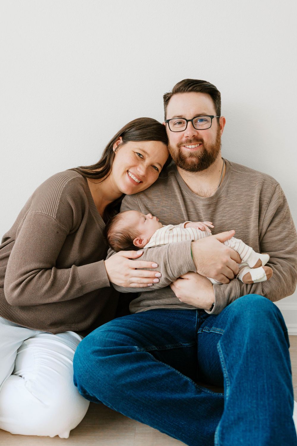 Smiling parents in neutral tones sit against white wall holding their sleeping newborn baby.