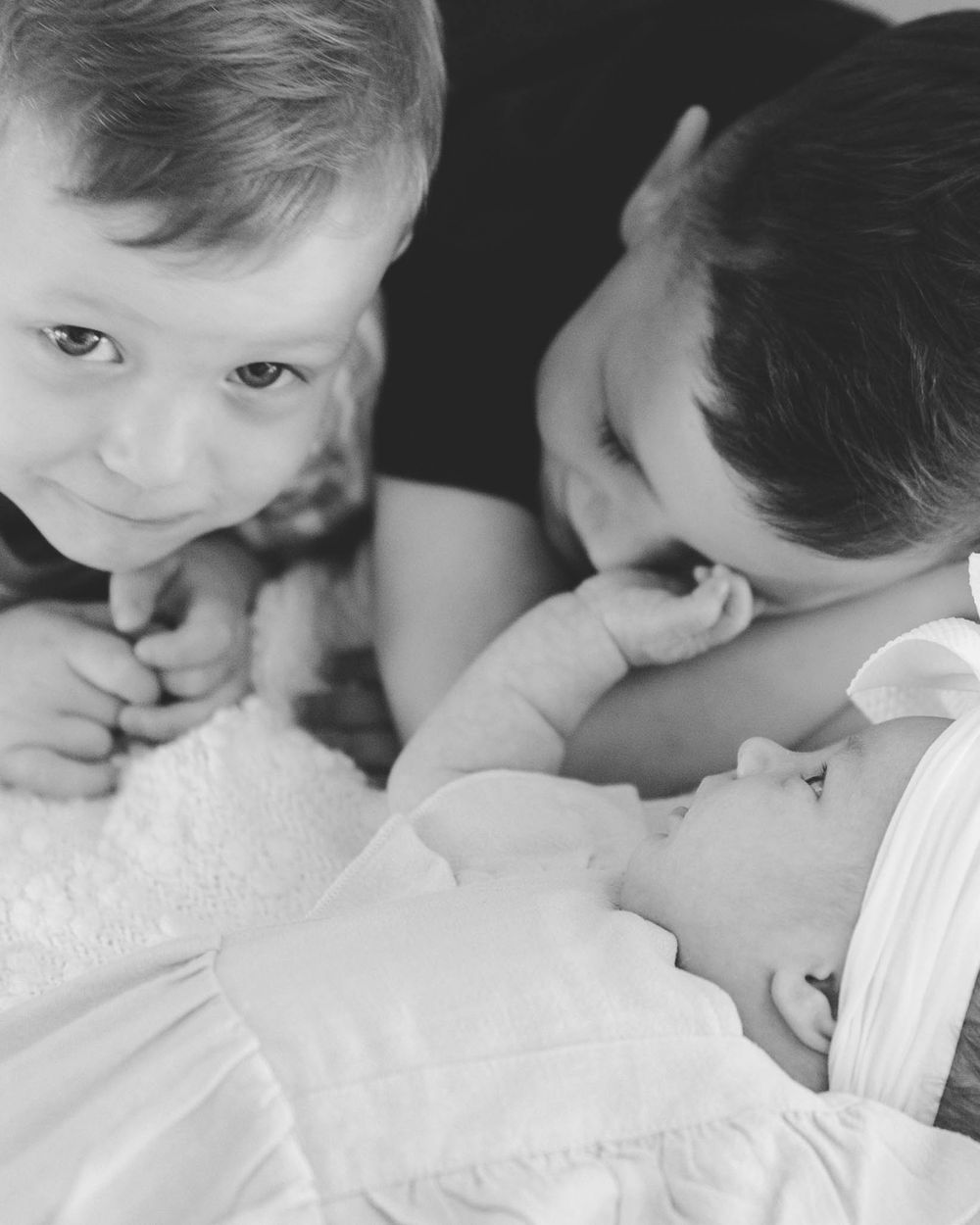 Two young siblings lean in close to gently interact with their sleeping newborn baby in black and white.
