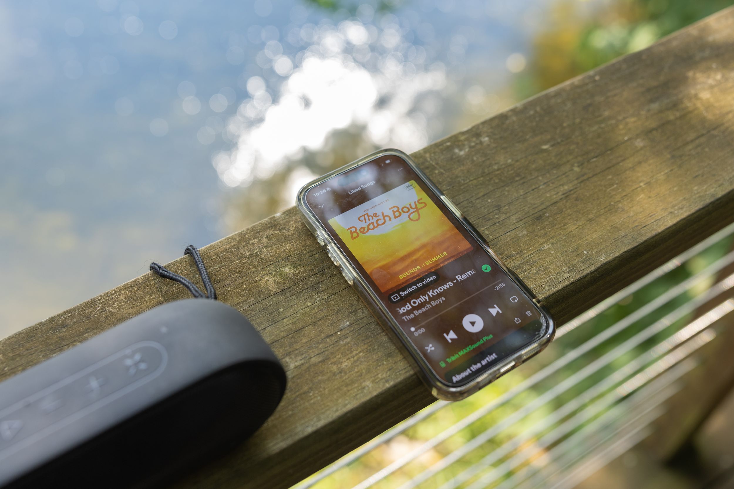 Smartphone displaying music app on wooden railing outdoors near water with Bluetooth speaker beside it.
