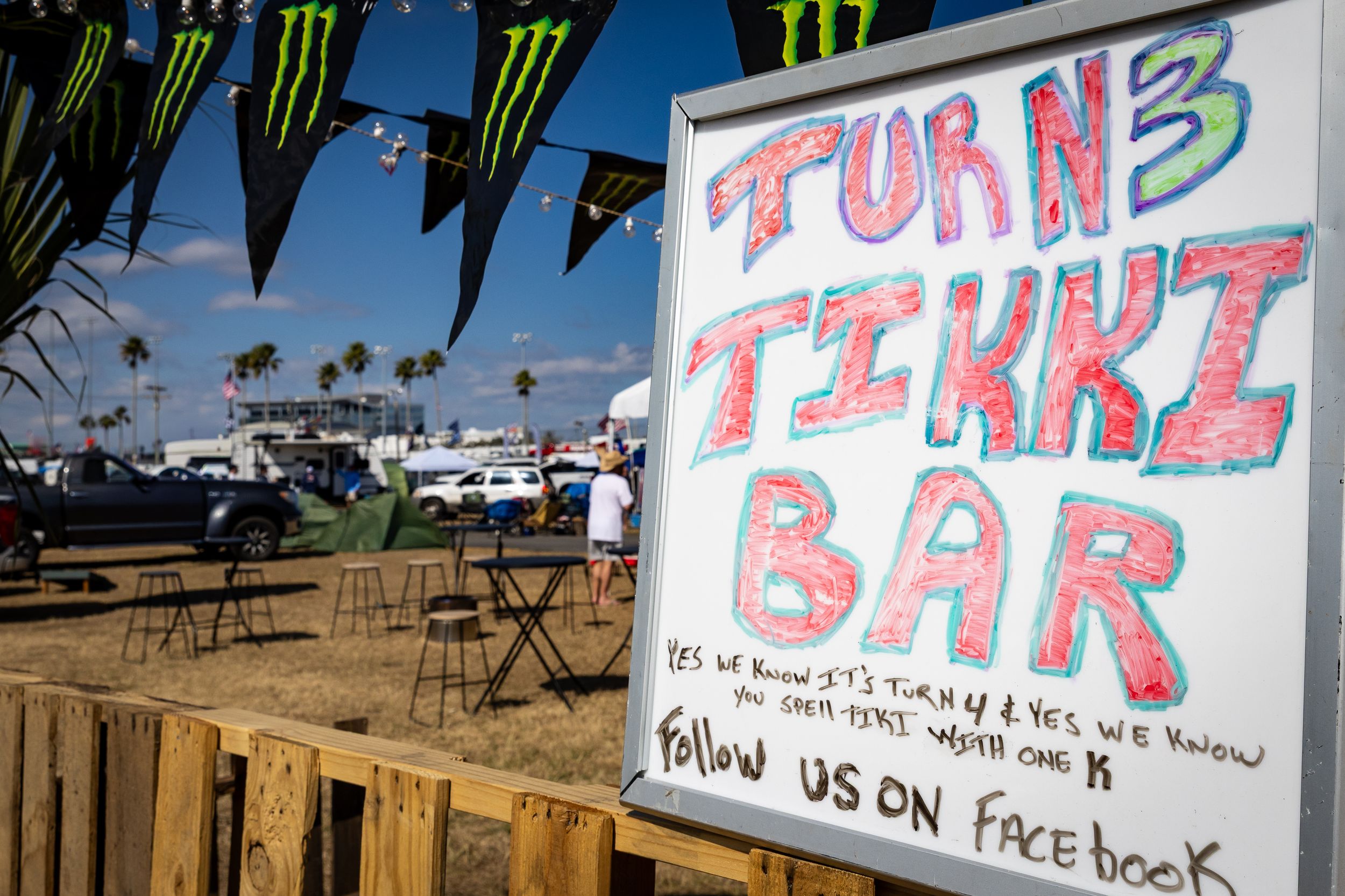 Colorful handmade sign for Turn 3 Tikki Bar at an outdoor racing event, inviting visitors to follow on Facebook.