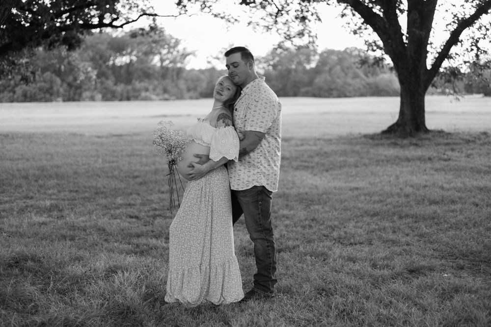 Expecting couple embracing beneath a large oak tree during an outdoor maternity photography session, captured in black and white.