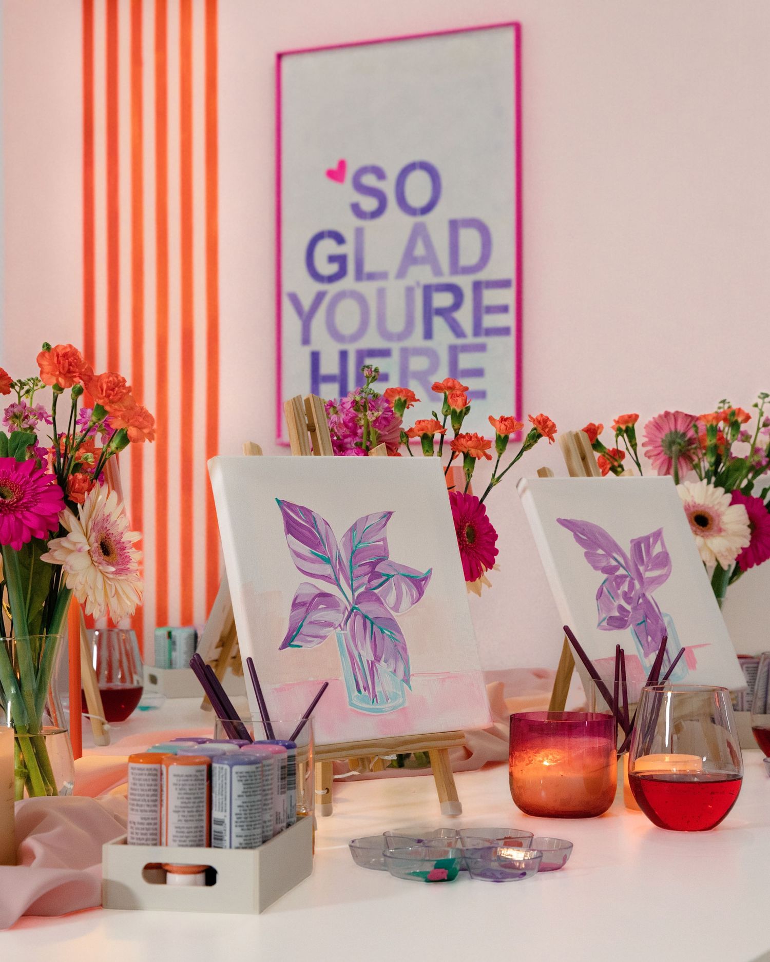 Woman in pink apron holding colorful items in a bright craft studio with art supplies in the background.