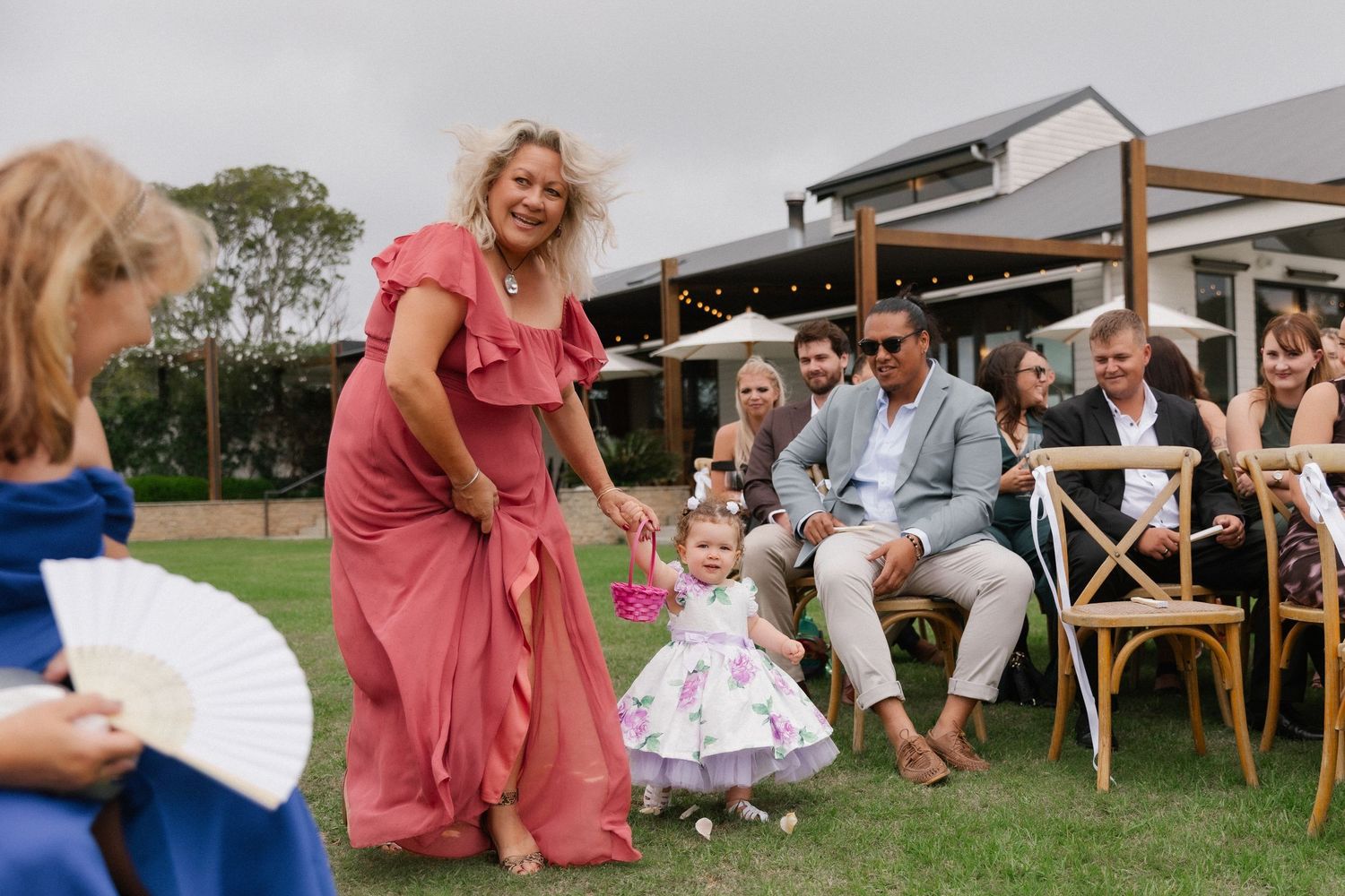 A woman in a pink dress guides a toddler flower girl down the aisle at an outdoor wedding ceremony.