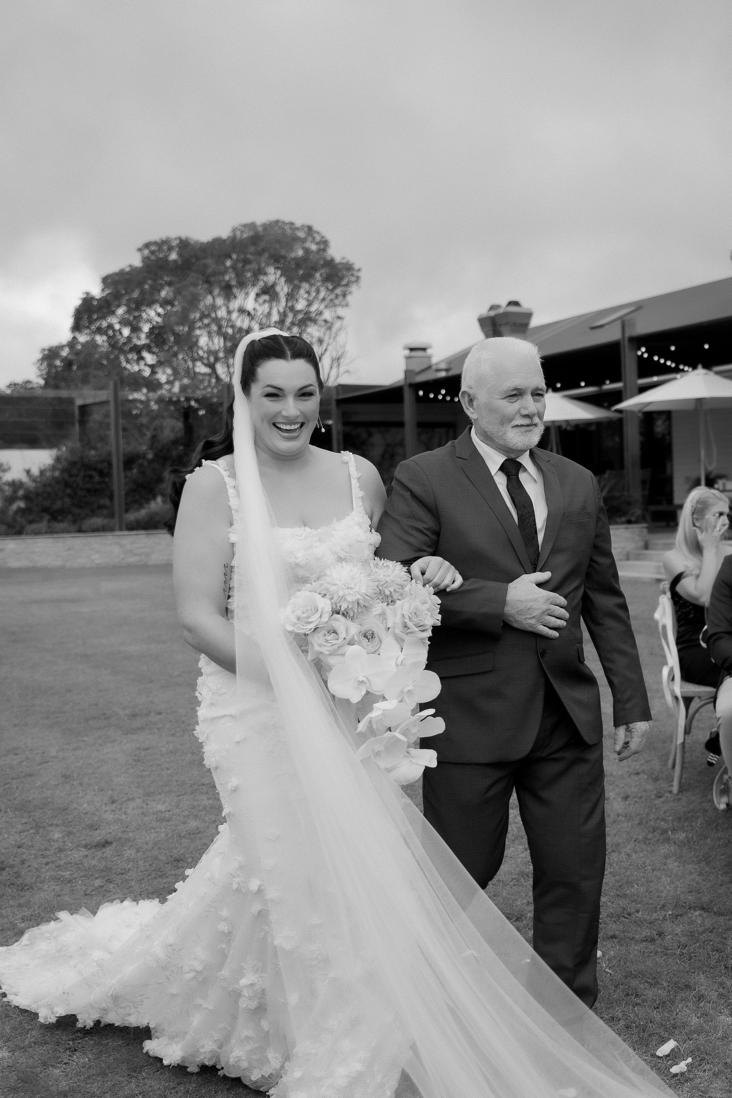 Black and white photo of smiling bride in floral gown walking down aisle with father on cloudy day.