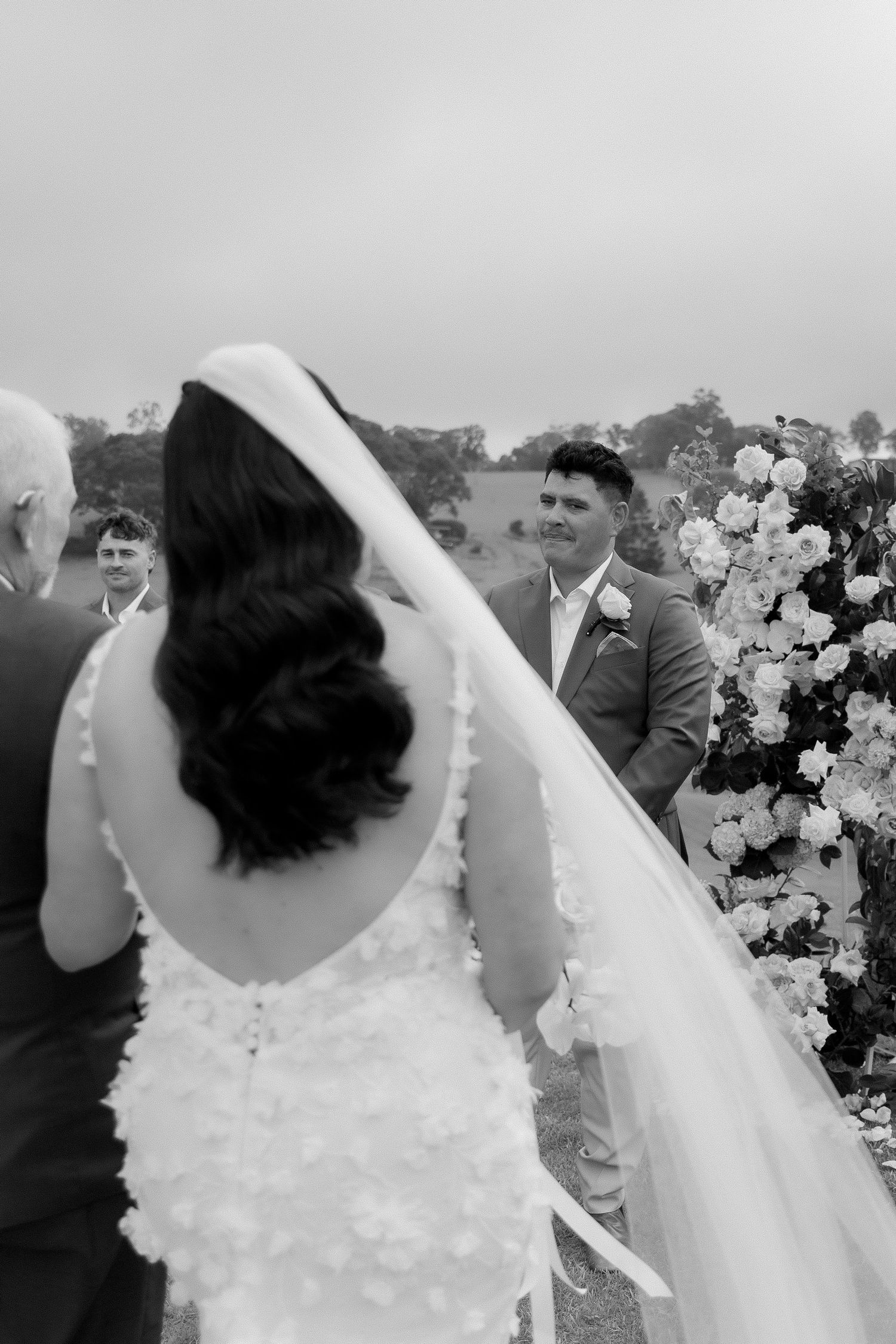 Black and white photo of a groom smiling as his bride walks toward him at outdoor wedding ceremony.