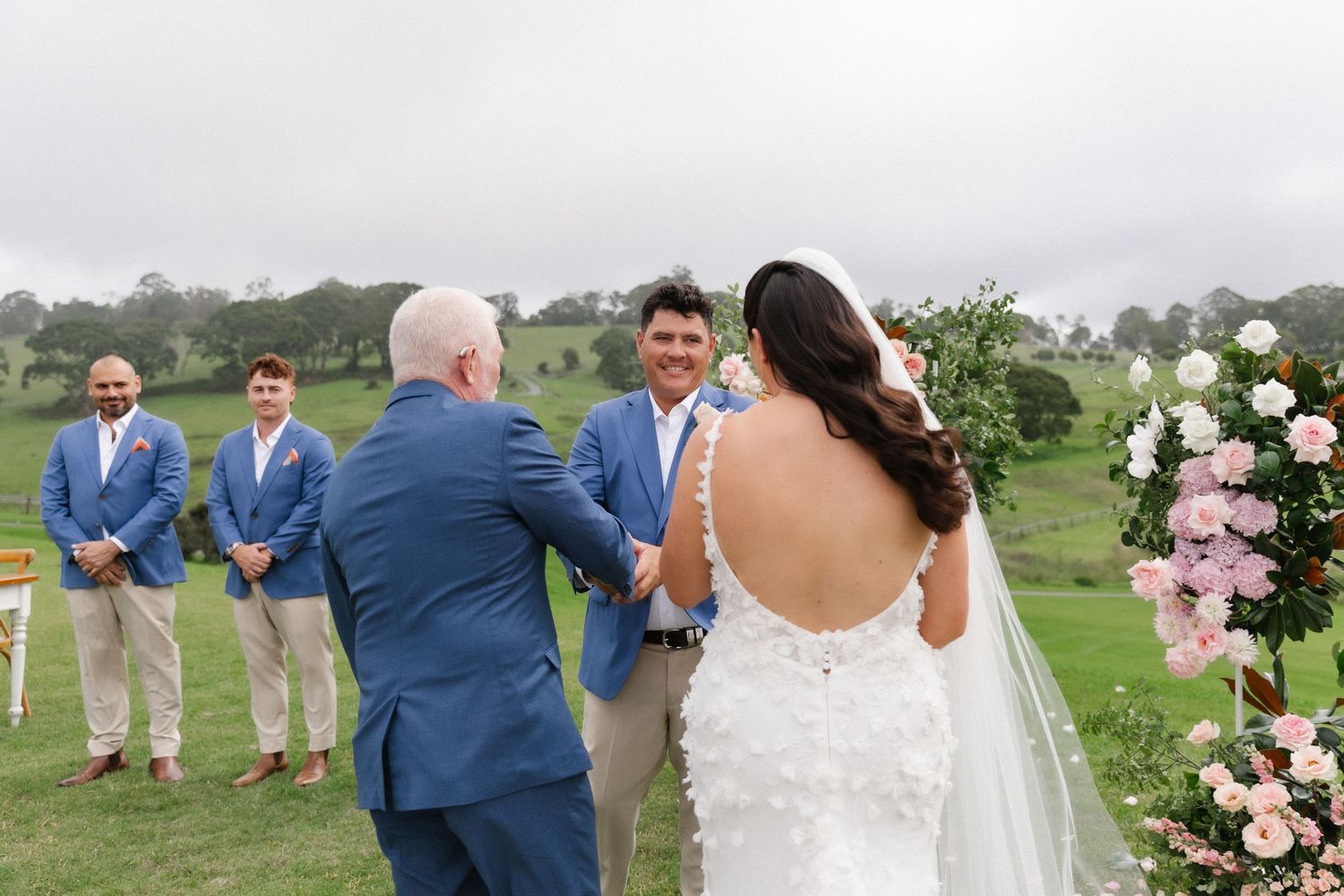 Bride and groom exchanging vows outdoors with groomsmen in blue suits at garden wedding.