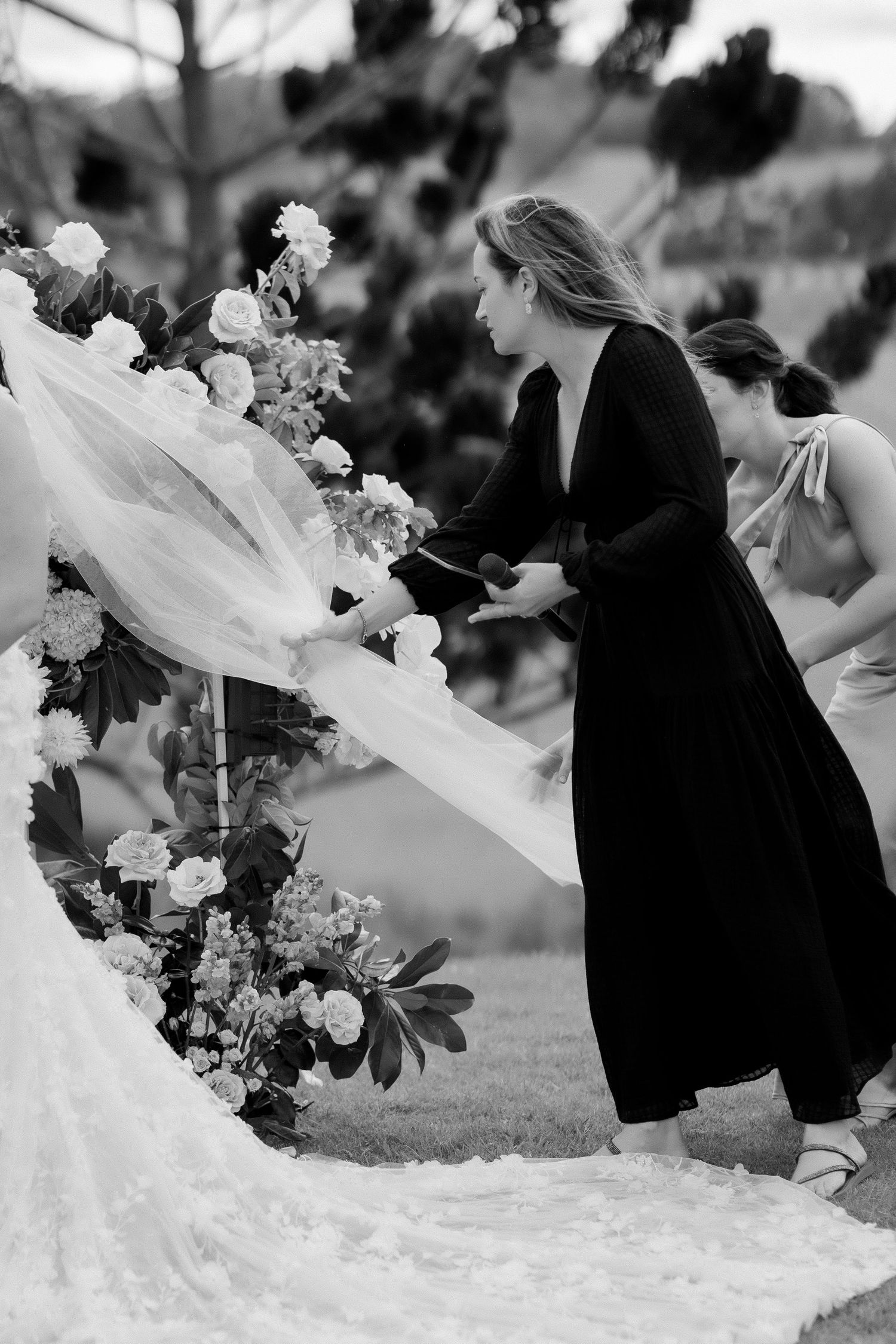 Black and white photo of a wedding coordinator arranging the brides veil near a floral arch outdoors.