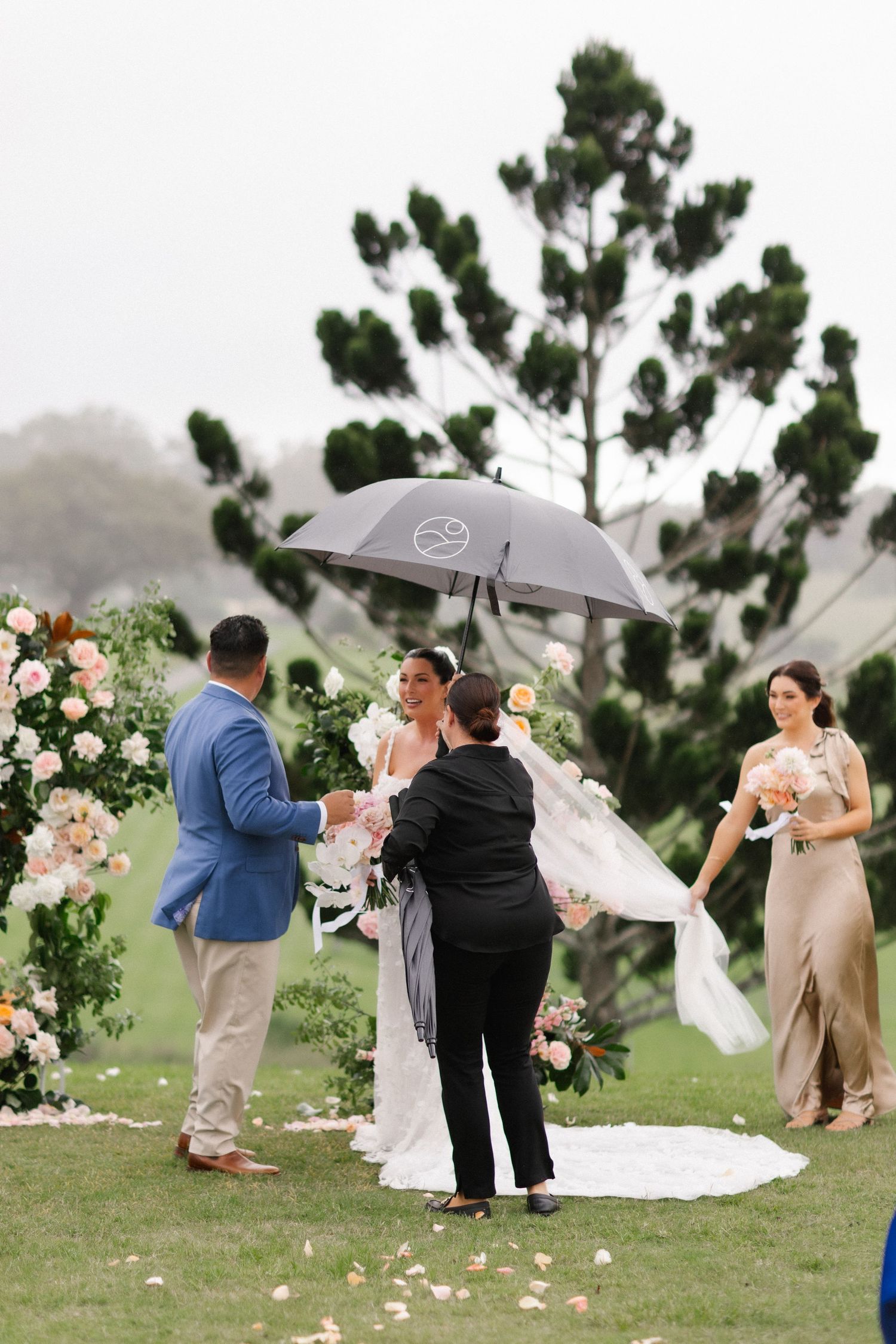 Outdoor wedding ceremony with bride and groom holding hands under umbrella, floral arch, bridesmaid, and lush green backdrop.