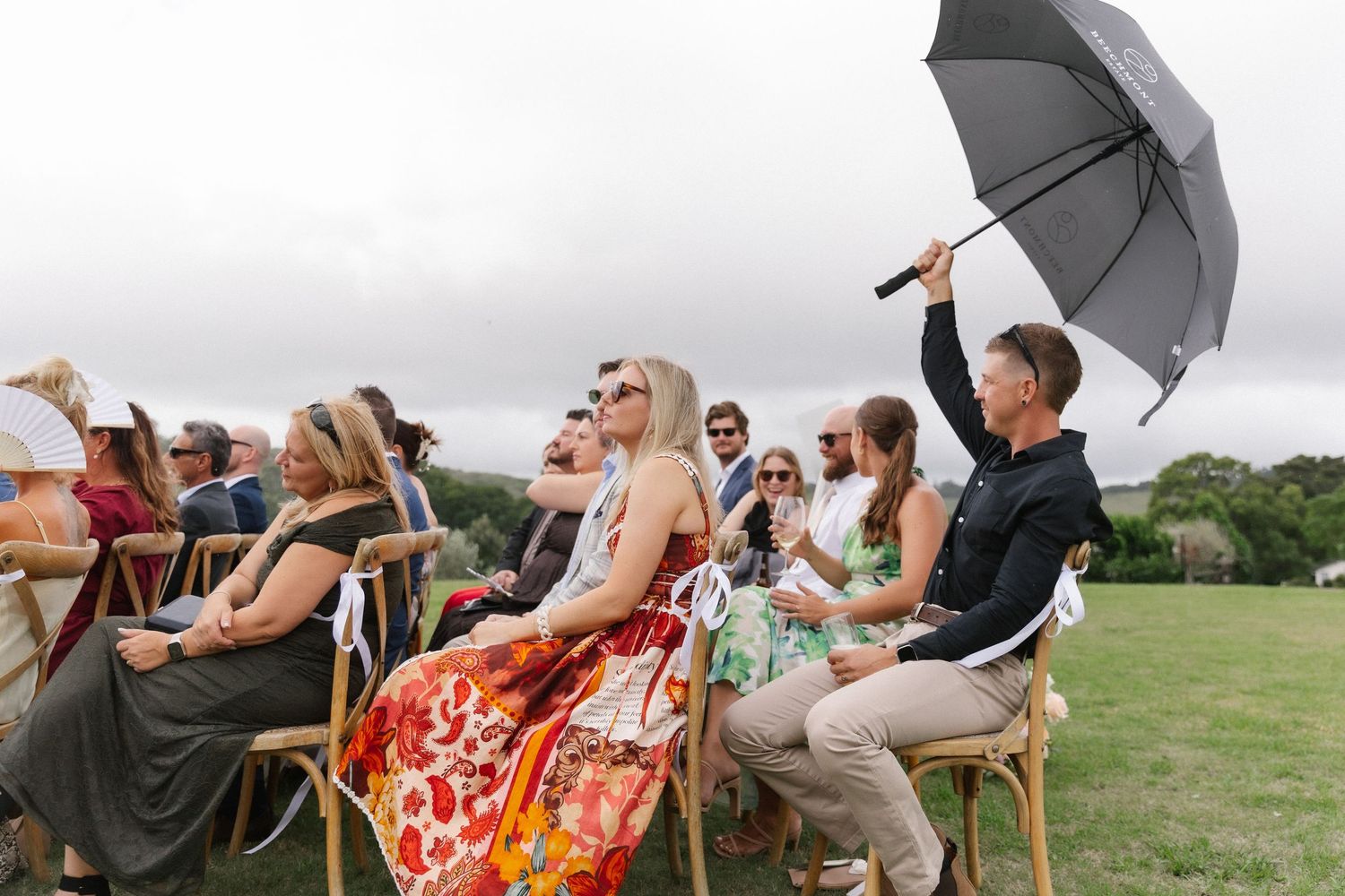 Wedding guests seated outdoors on a cloudy day, one holding a black umbrella for shade.