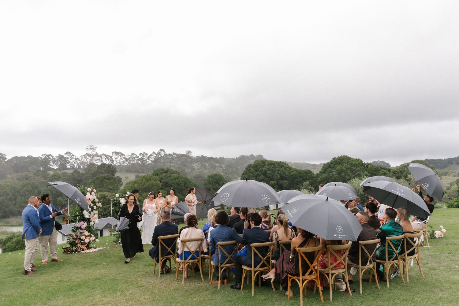 Wedding ceremony on green lawn with guests holding clear umbrellas under overcast skies.