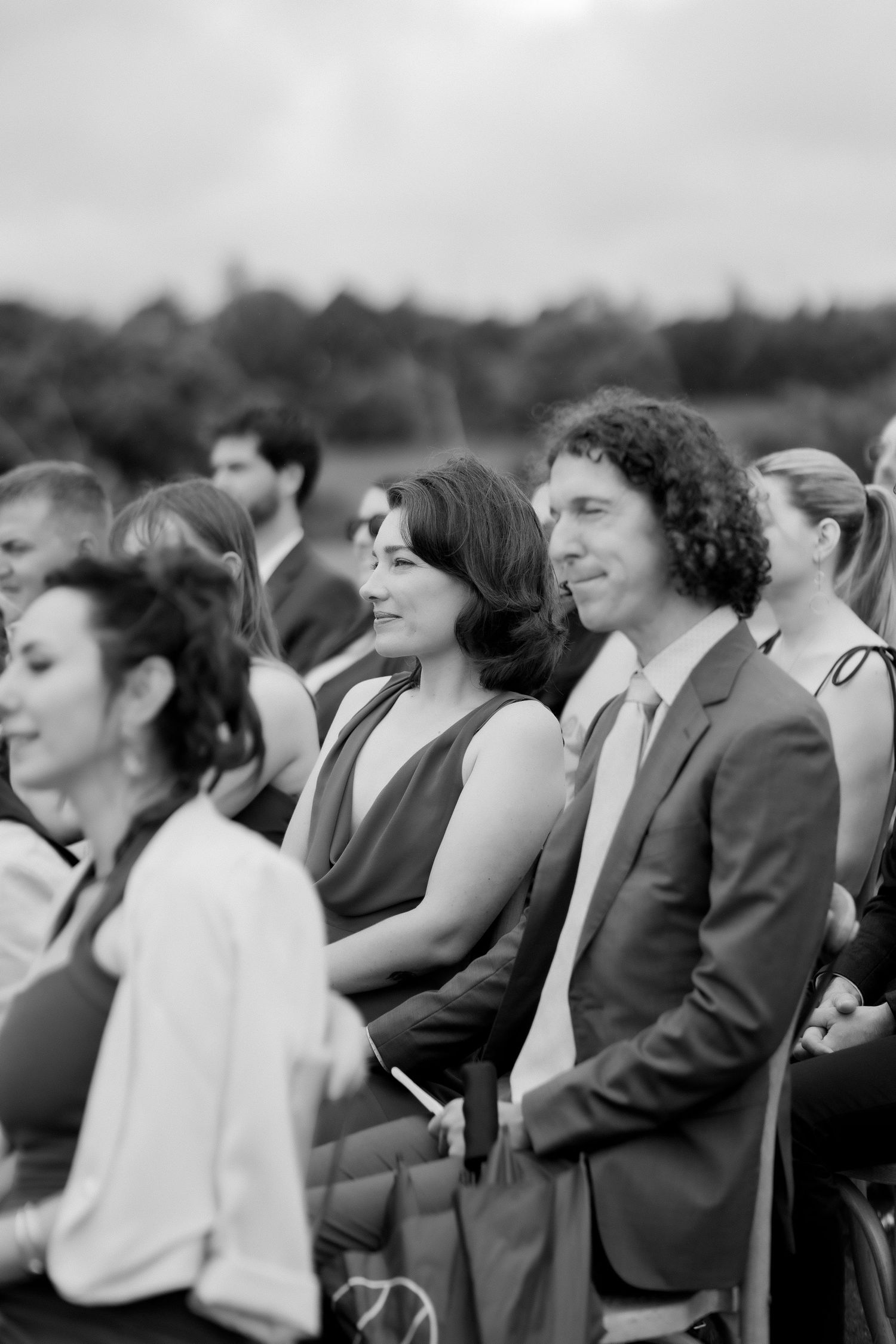 Black and white photo of wedding guests seated outdoors during a ceremony, smiling couple in foreground.