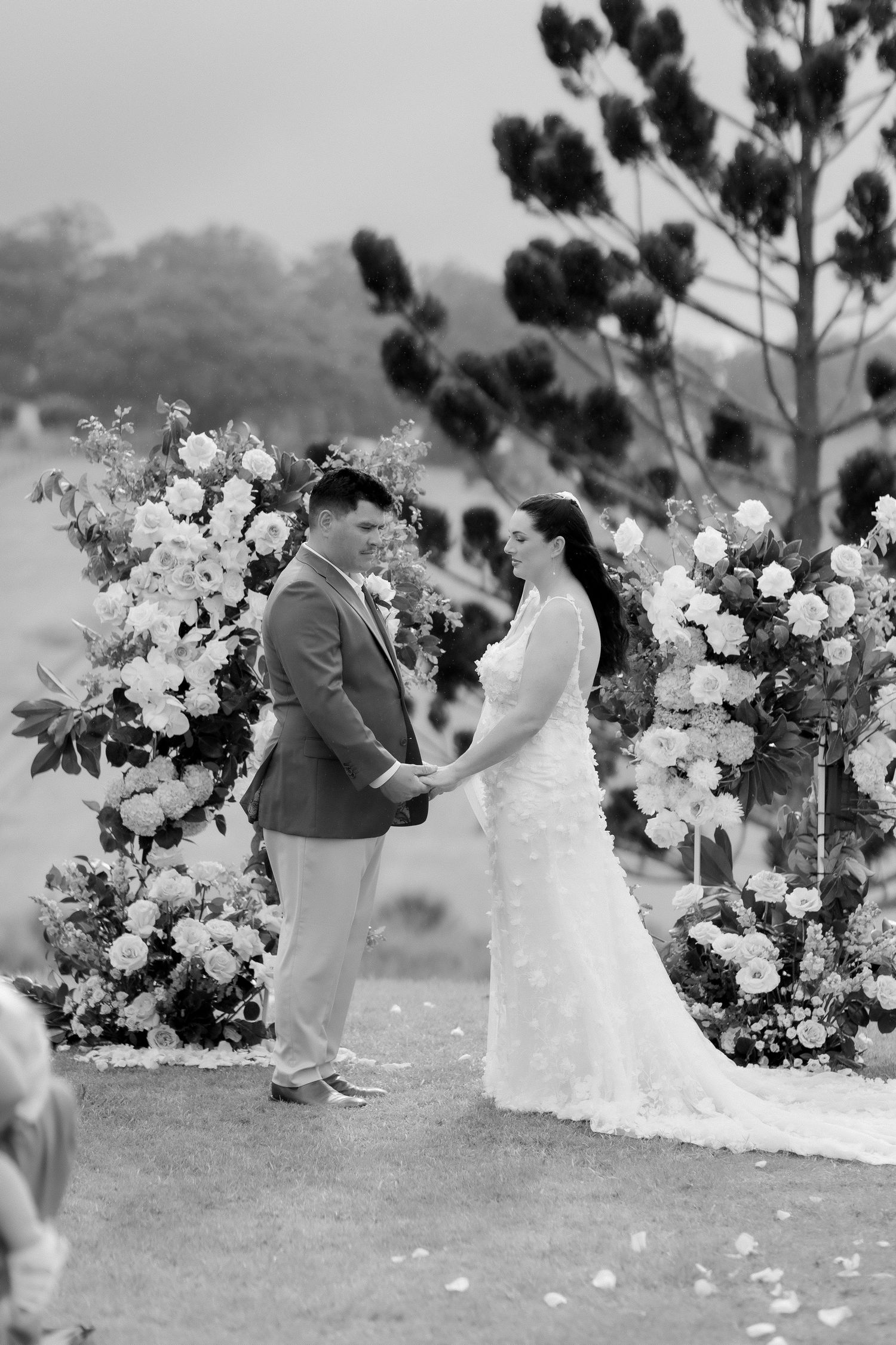 Black and white photo of bride and groom exchanging vows at a floral arch ceremony outdoors.