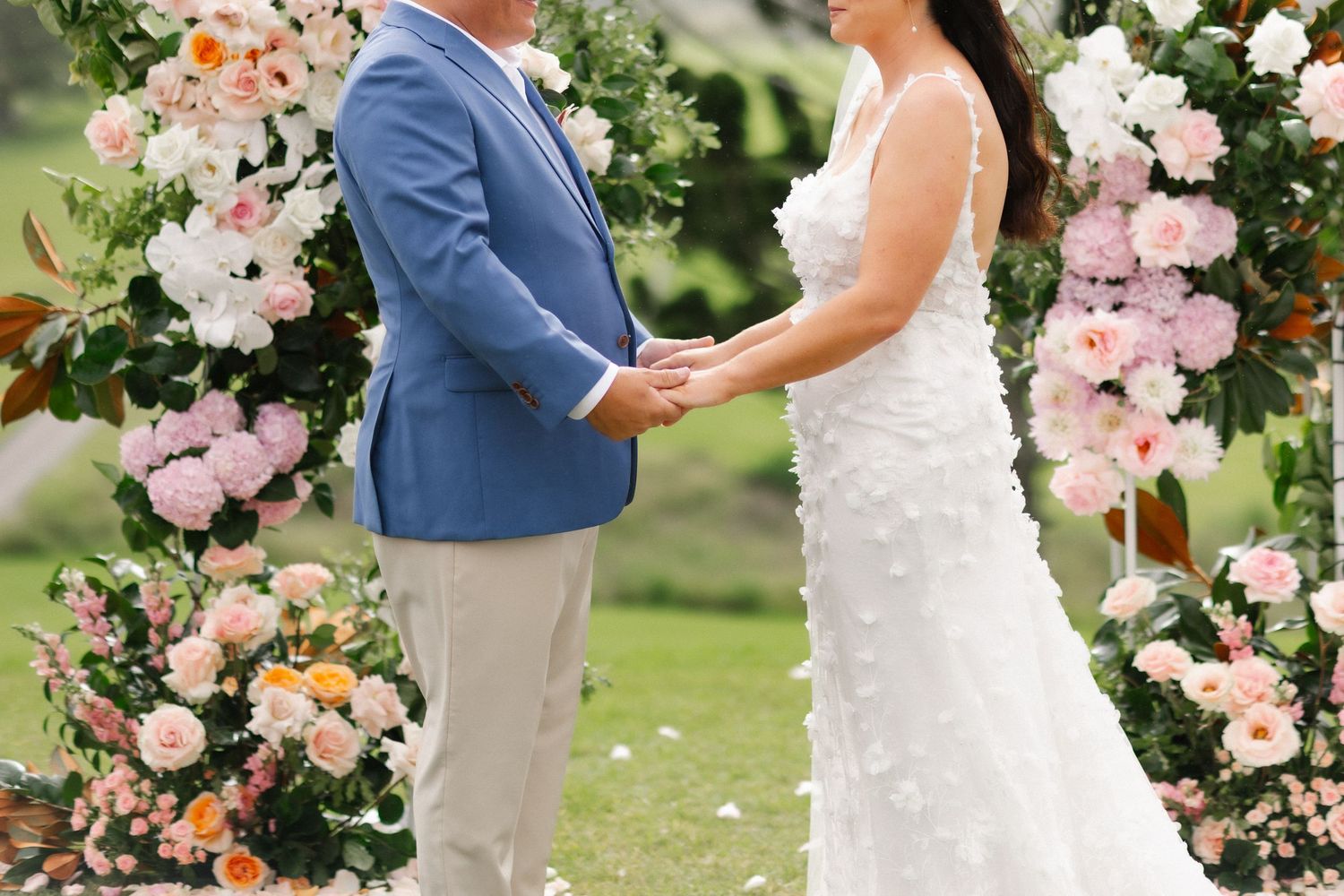 Bride and groom holding hands at floral arch adorned with pink and white roses during ceremony.