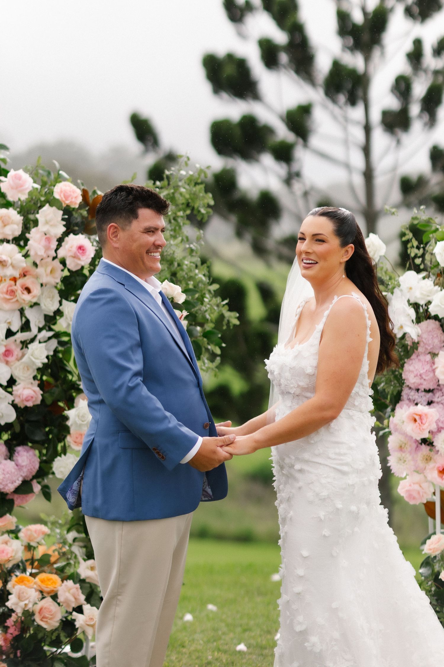 Smiling bride and groom holding hands at outdoor wedding ceremony beside a pink and white floral arch.