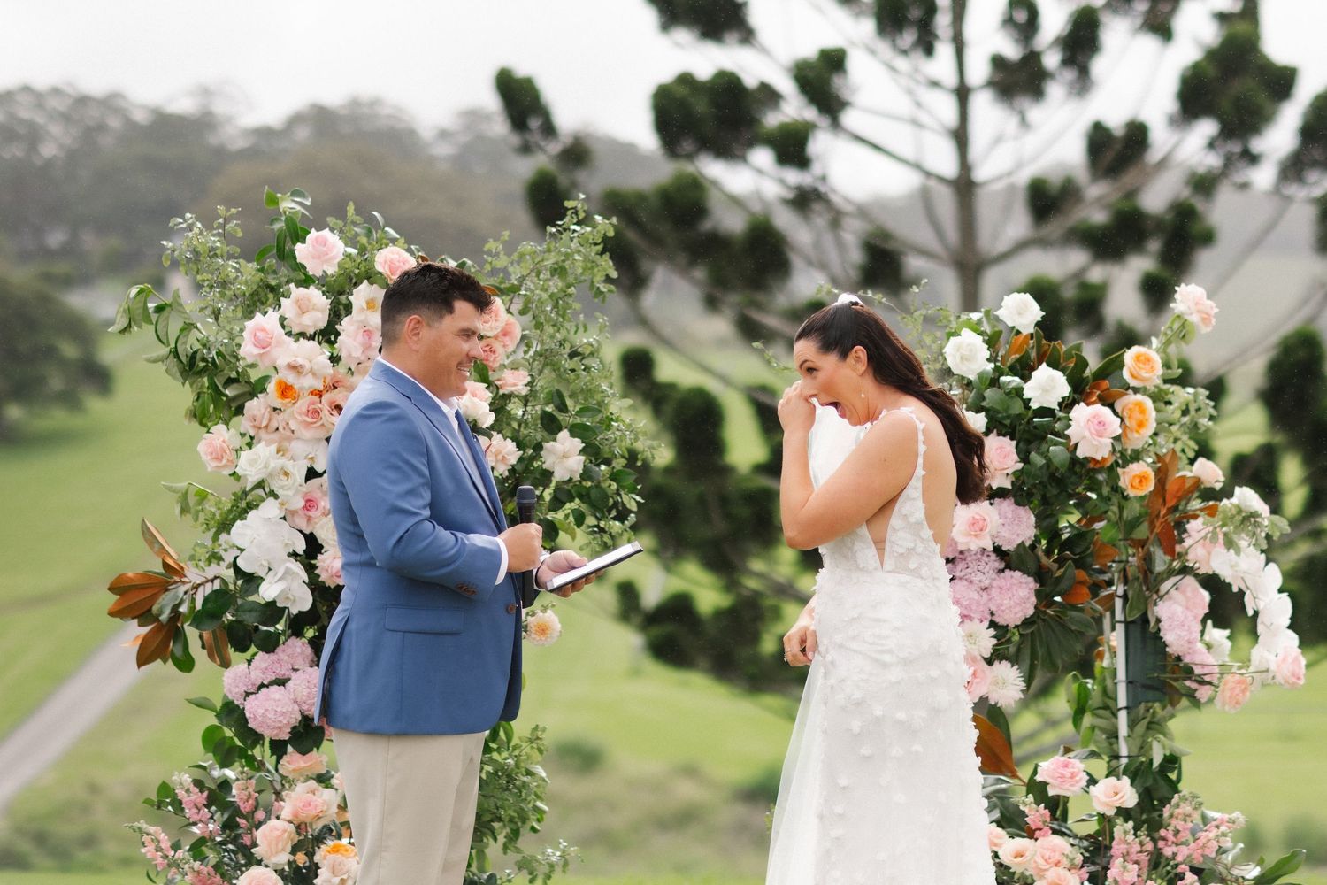 Groom in blue blazer reading vows as emotional bride stands before a lush floral arch outdoors.