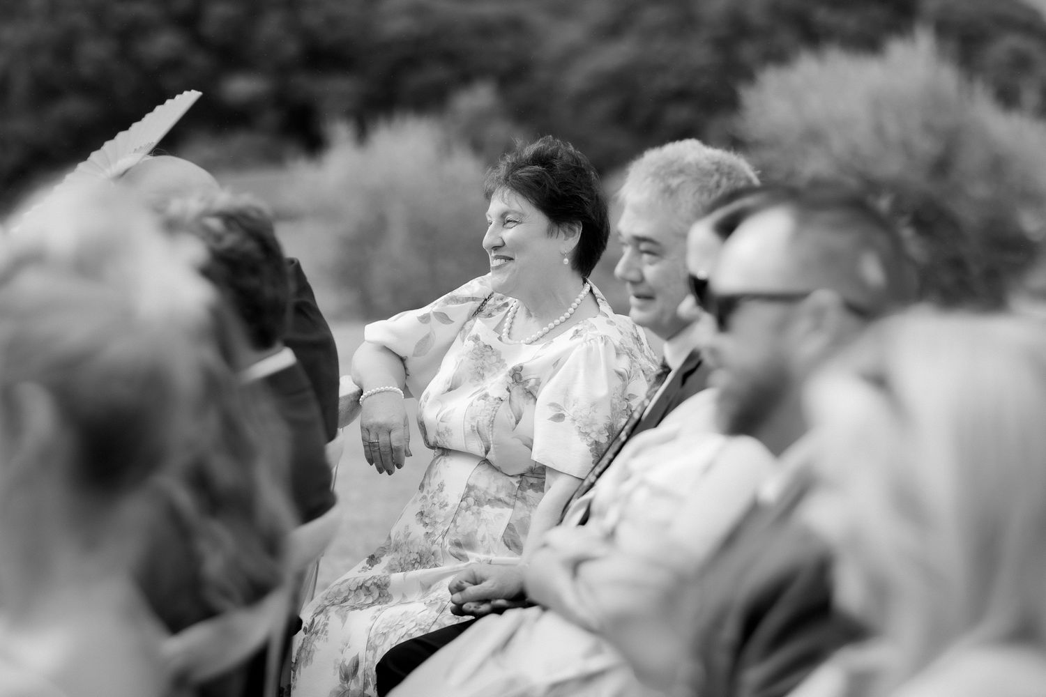 Black and white candid photo of smiling wedding guests seated outdoors during wedding ceremony.