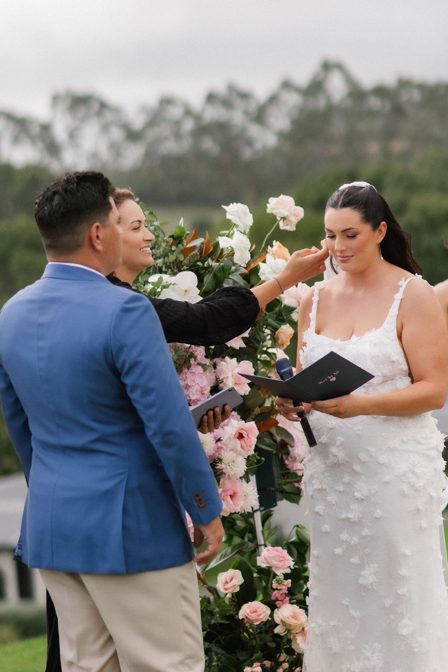 Color photo of a celebrant touching the brides face during an outdoor wedding ceremony with floral arch.