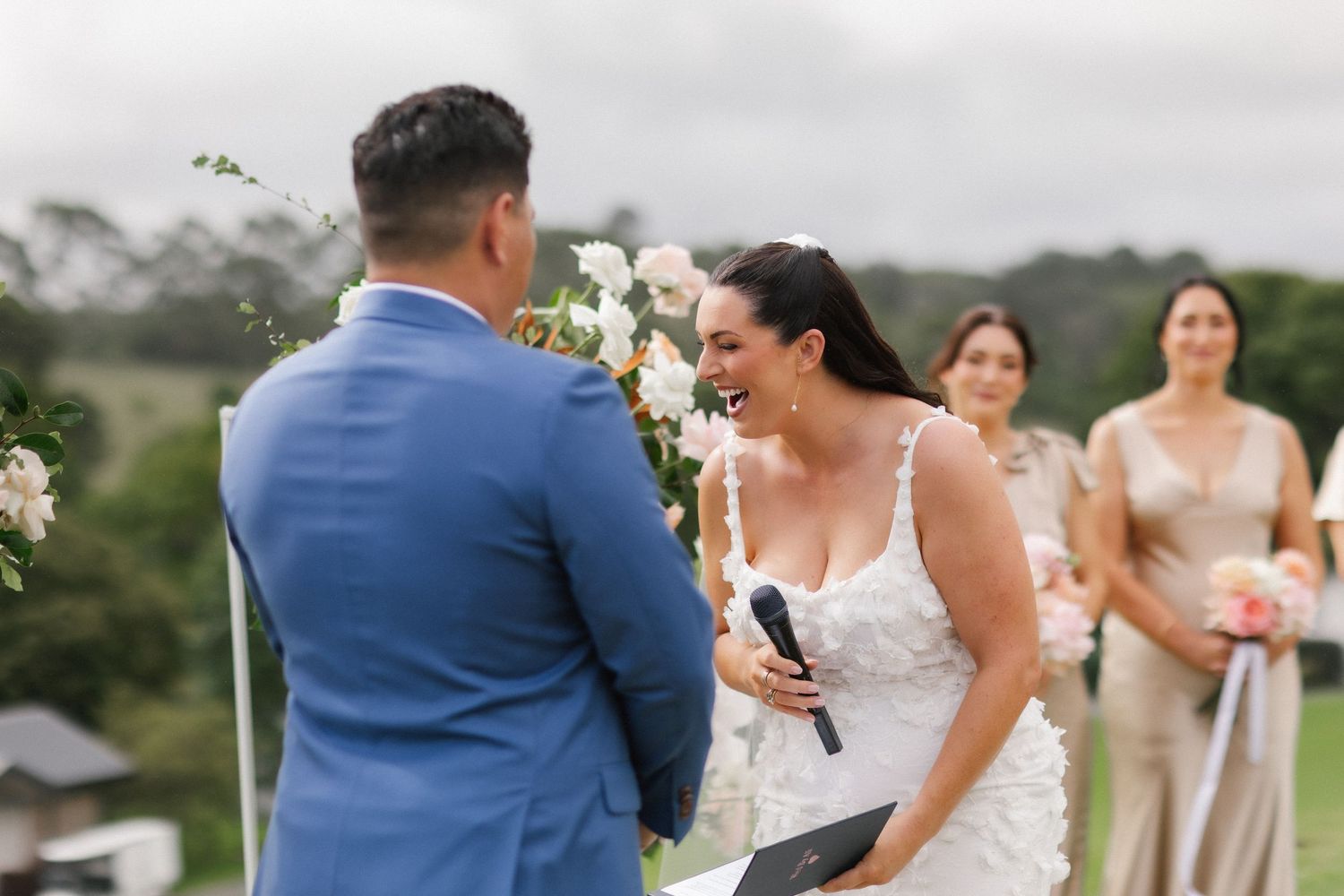 Bride in white gown laughs joyfully with groom in blue suit during outdoor wedding ceremony with floral arch.