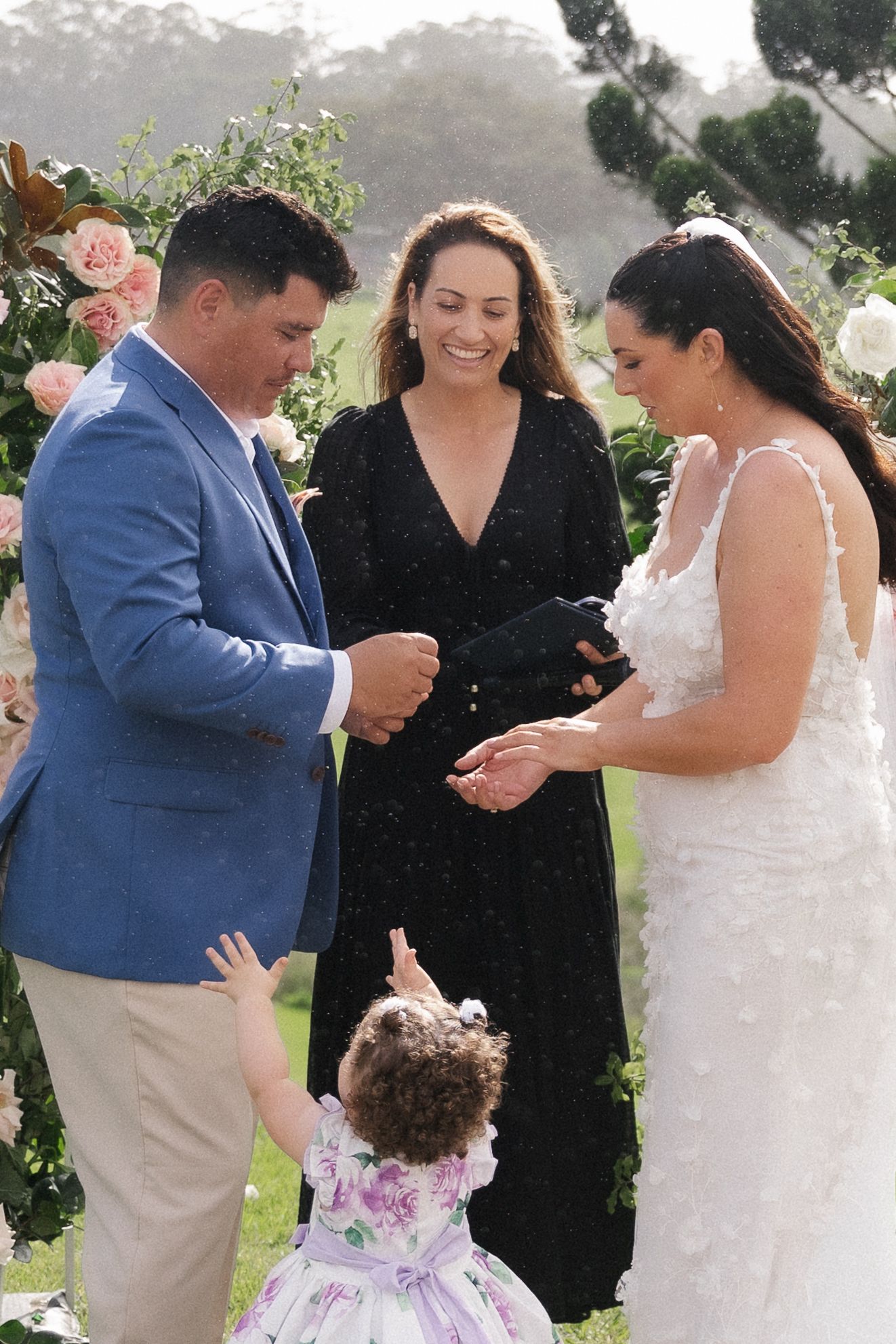 Bride and groom exchanging rings at outdoor wedding ceremony with celebrant and flower girl.