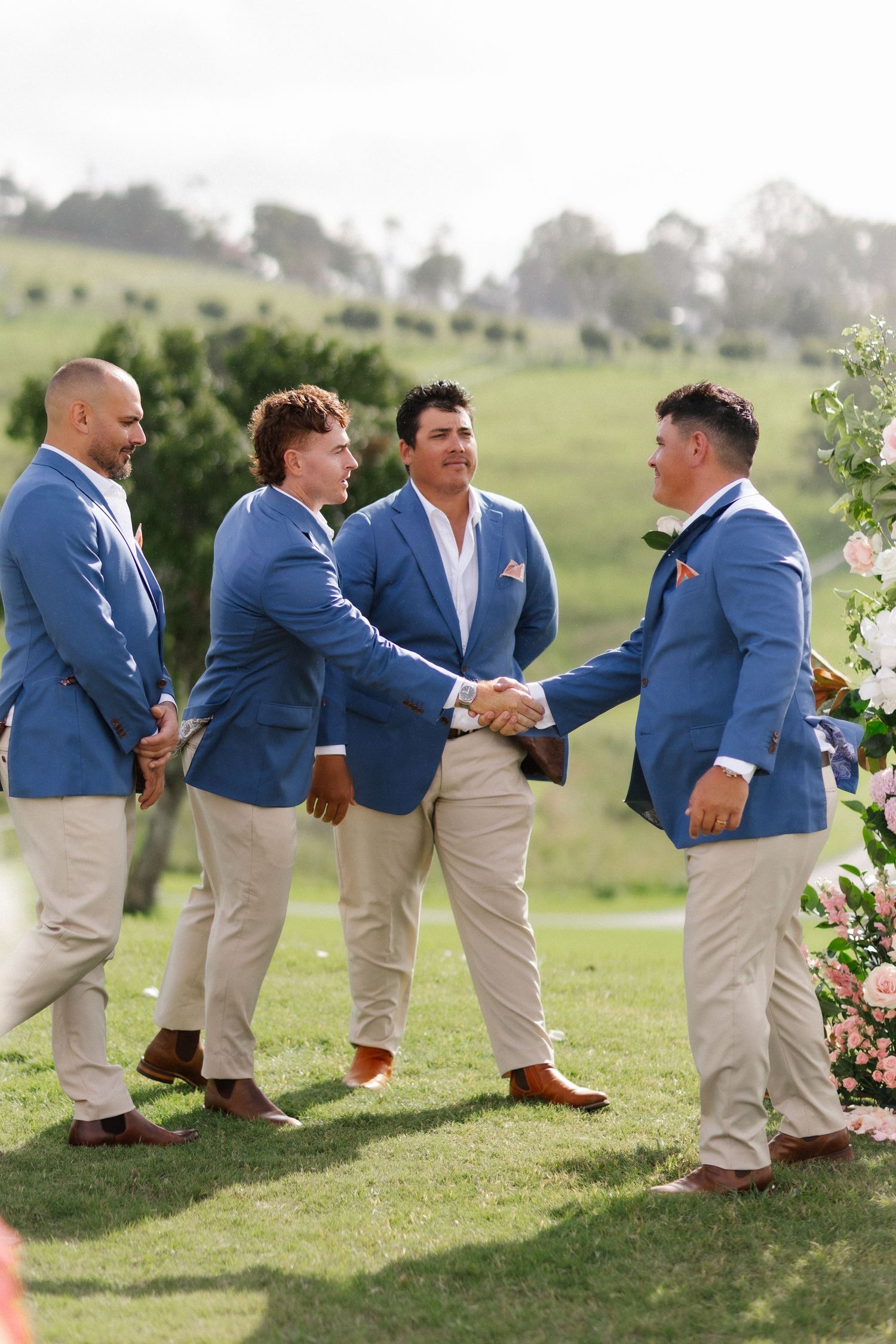 Groomsmen in matching blue blazers and khaki pants shaking hands near floral arch on green lawn.