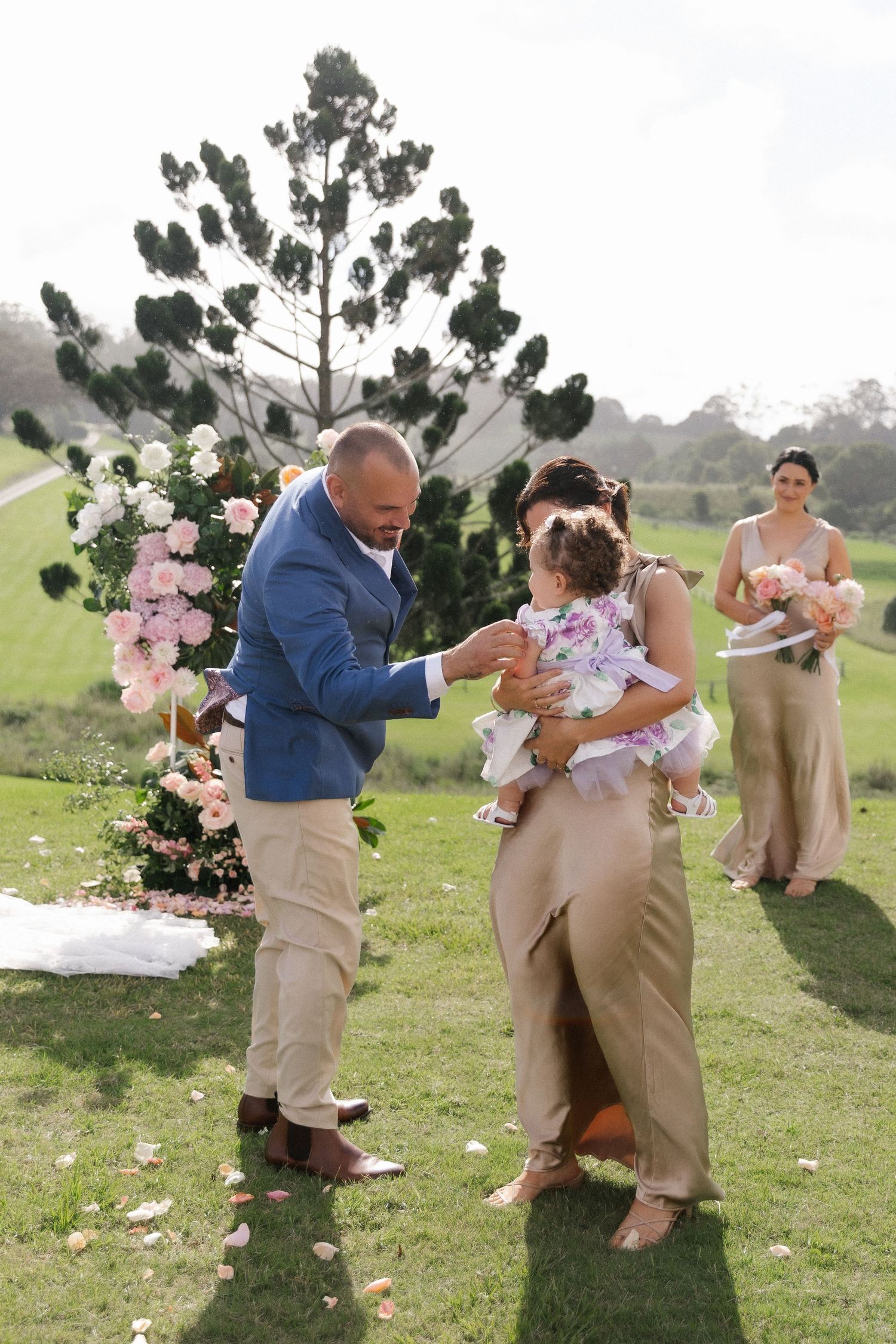 Groom in blue blazer and woman in gold dress interact with a baby girl during outdoor wedding ceremony.
