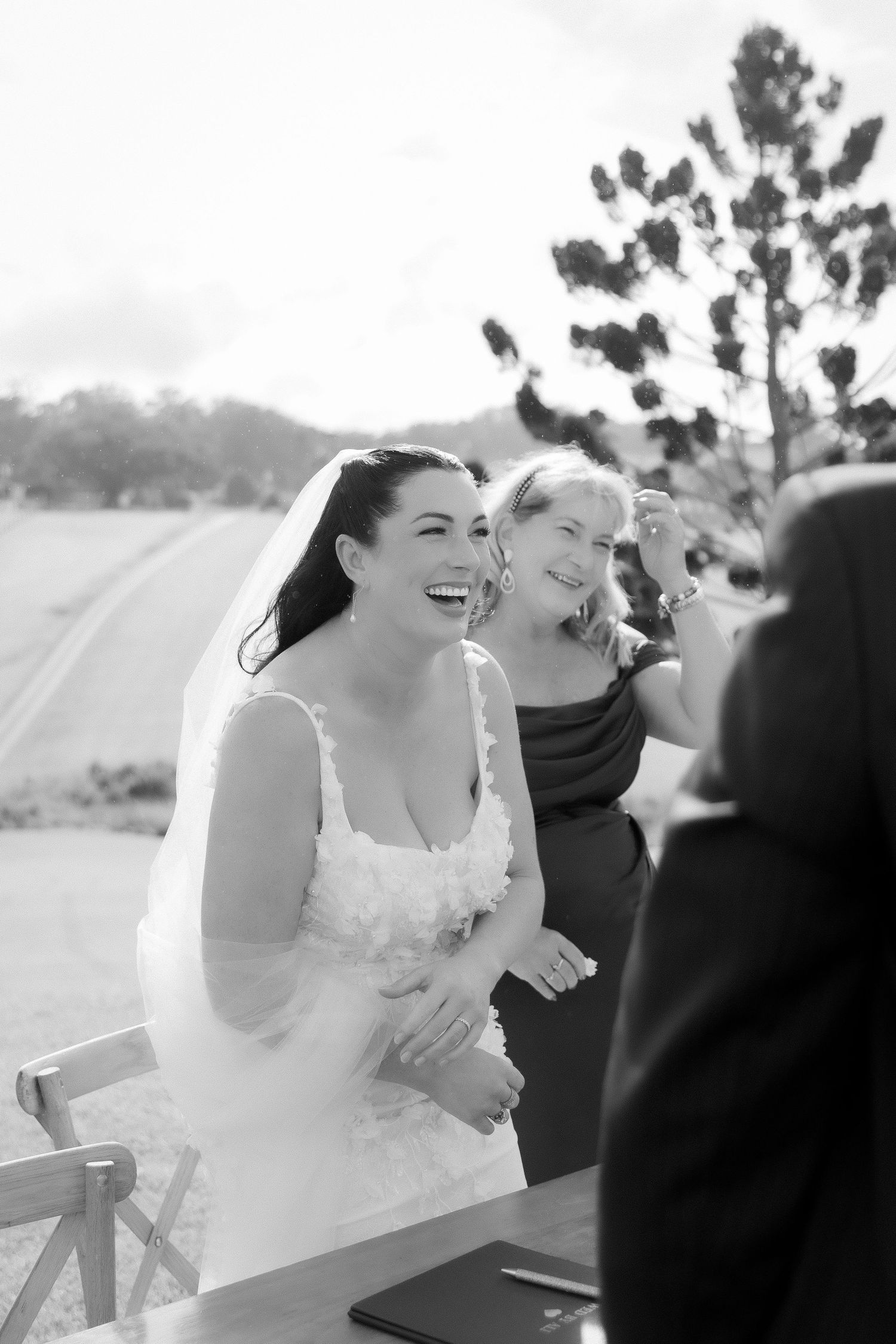 Black and white photo of laughing bride in veil with bridesmaid during outdoor wedding ceremony.