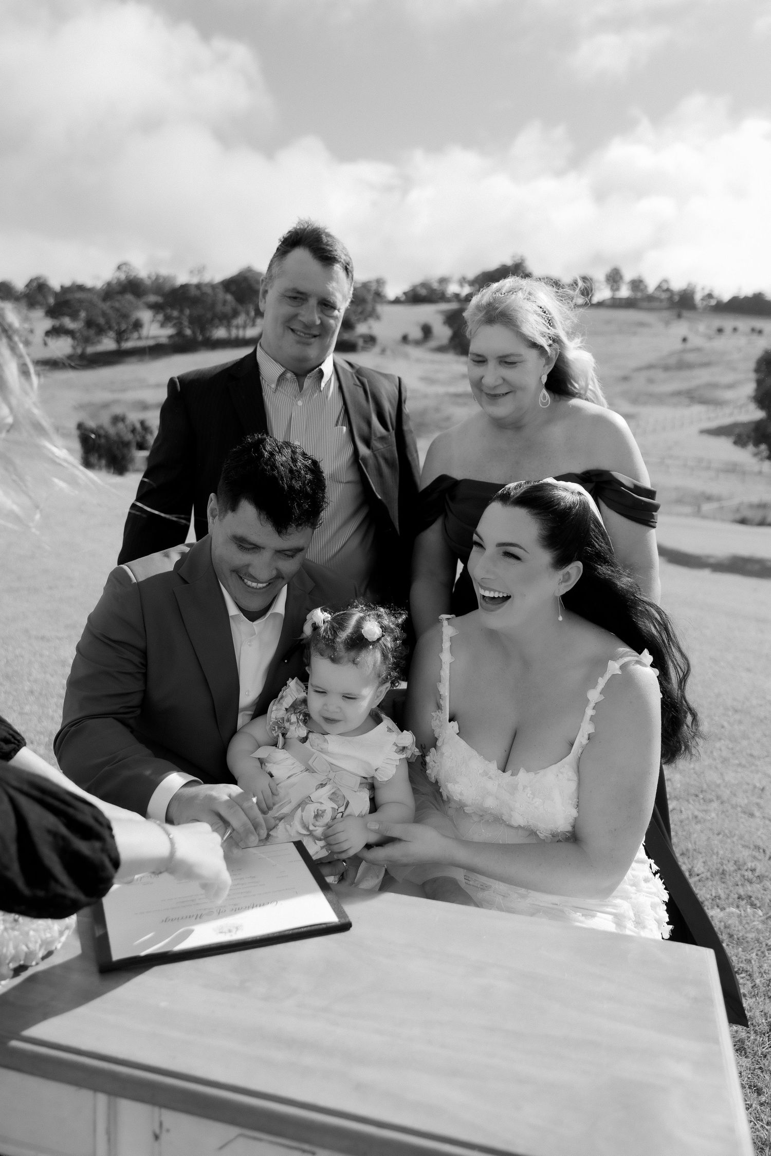 Black and white photo of bride and groom signing wedding register with witnesses and flower girl at outdoor ceremony.
