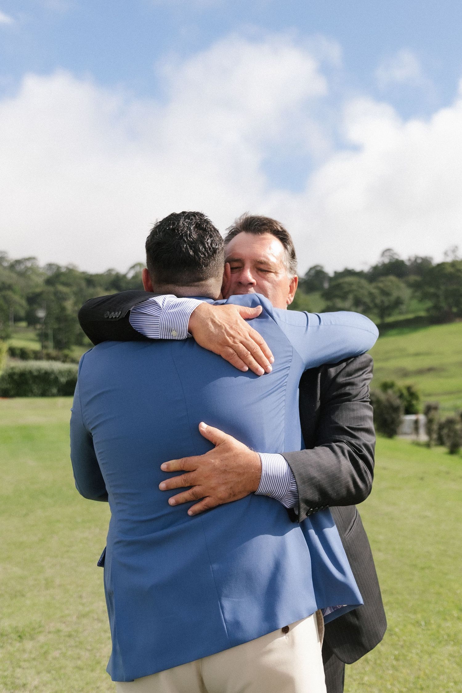Groom in blue suit sharing warm embrace with older man in gray suit at outdoor wedding reception.