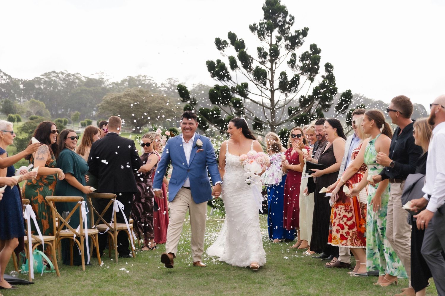 Newlywed couple walking down aisle as guests throw white confetti at outdoor garden wedding.