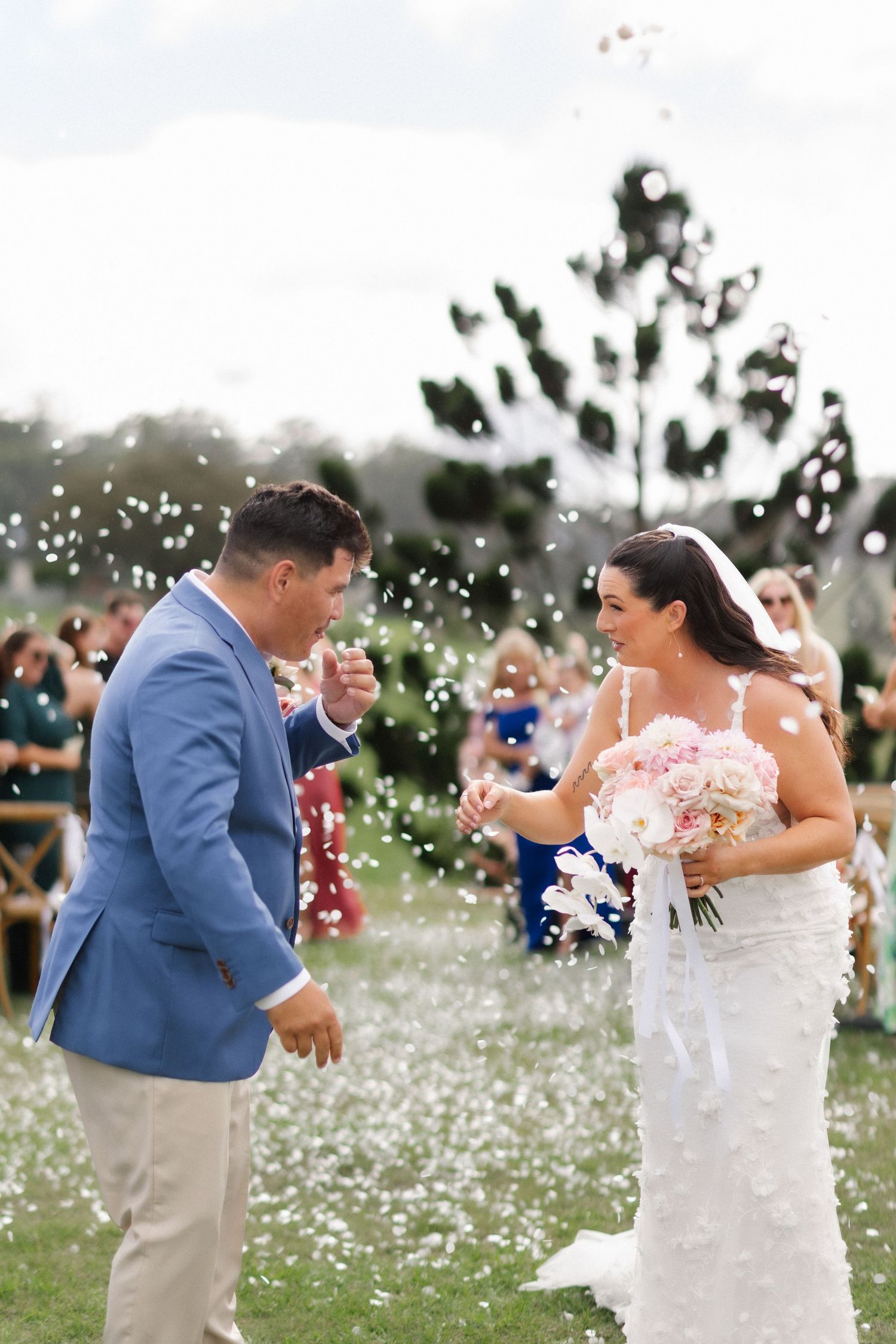 Bride and groom laughing as guests shower them with white flower petals during outdoor ceremony exit.