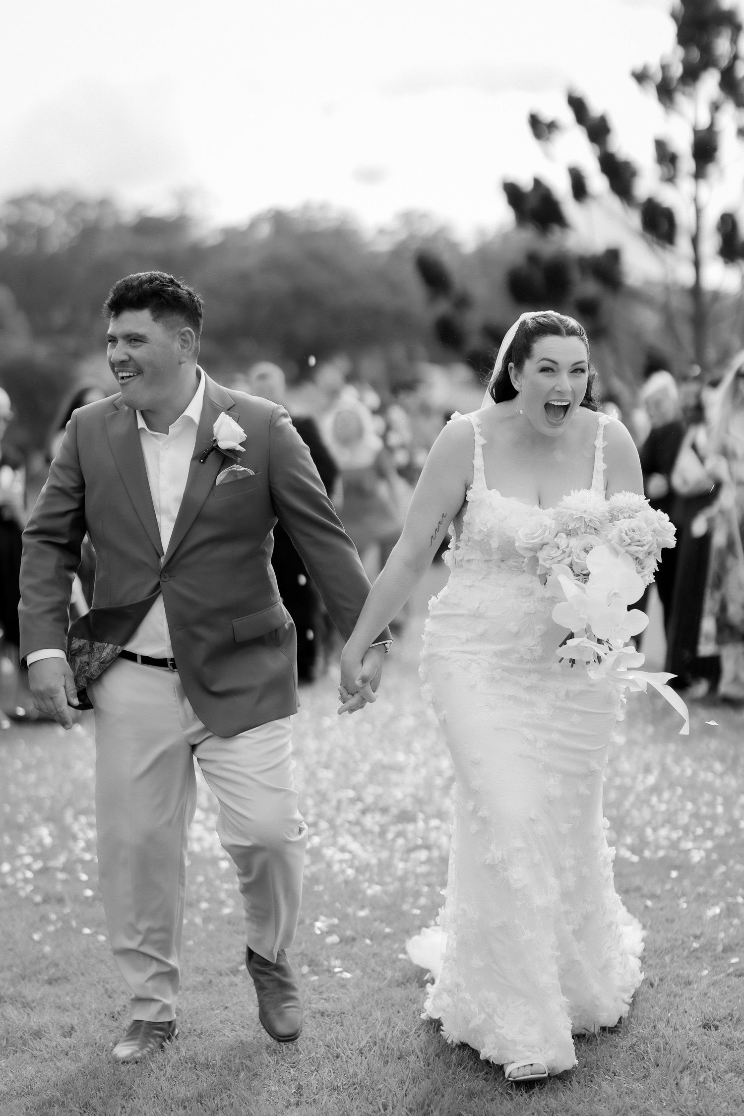 Black and white photo of joyful bride and groom walking through confetti after outdoor wedding ceremony.