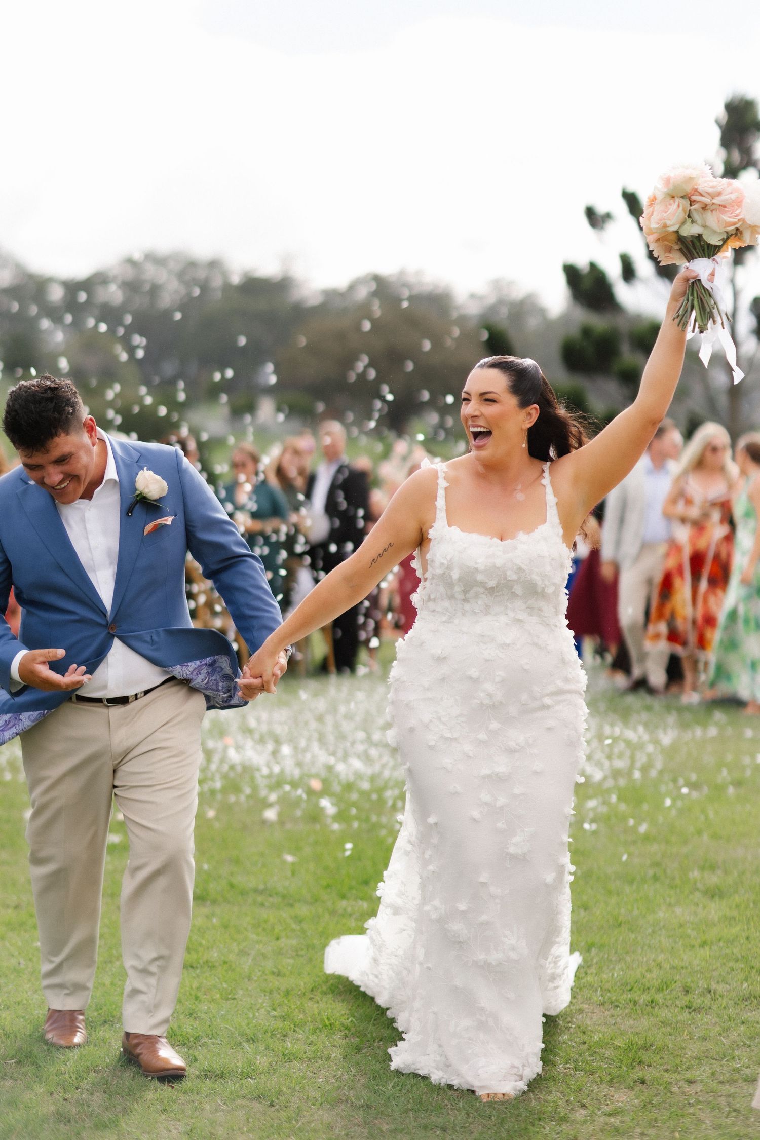 Joyful bride in white floral gown raises bouquet while holding groom's hand during outdoor wedding reception exit.