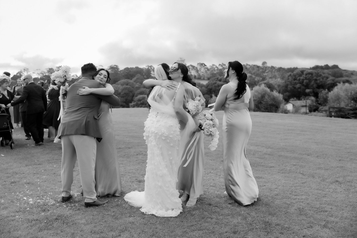 Black and white photo of bride in lace gown embracing wedding party on green lawn.