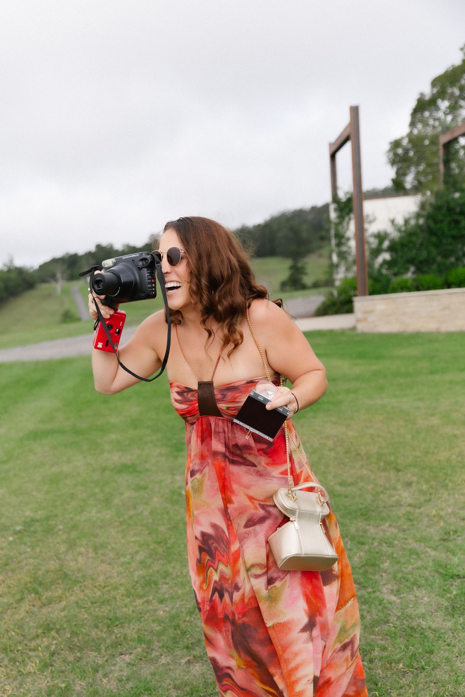 Woman in floral maxi dress laughing while taking photos with DSLR camera outdoors.