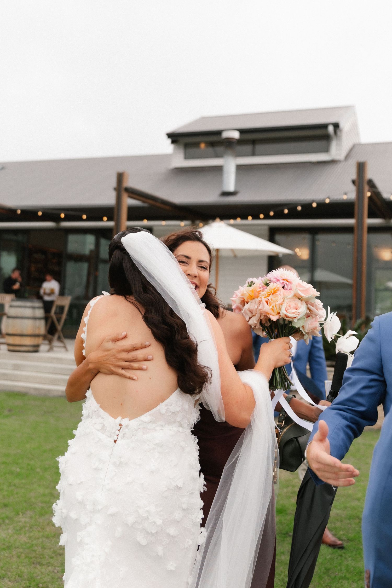 Bride in white lace gown hugging bridesmaid in brown dress holding pink bouquet at outdoor wedding venue.