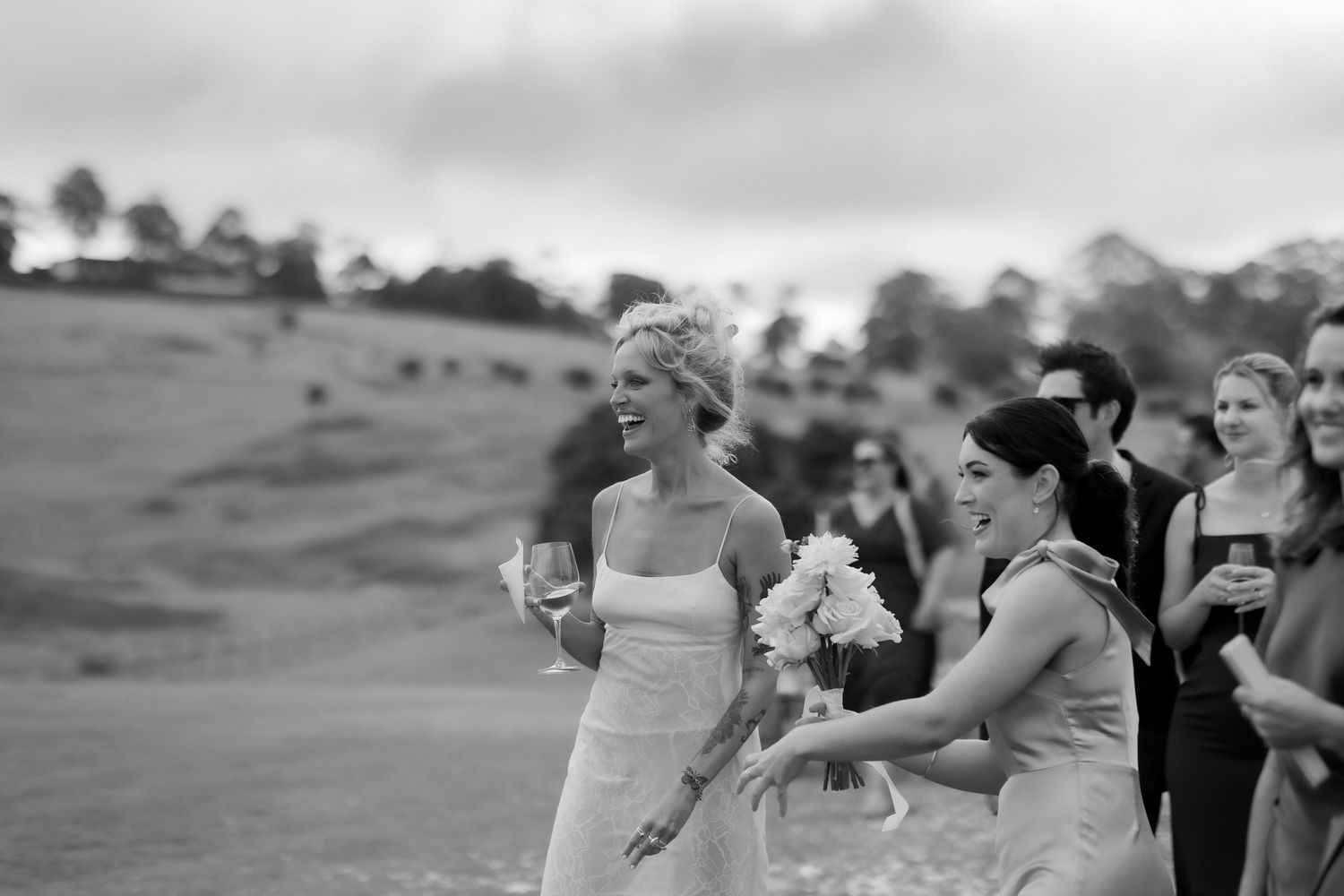 Black and white photo of bride and bridesmaid laughing outdoors at wedding reception in countryside.