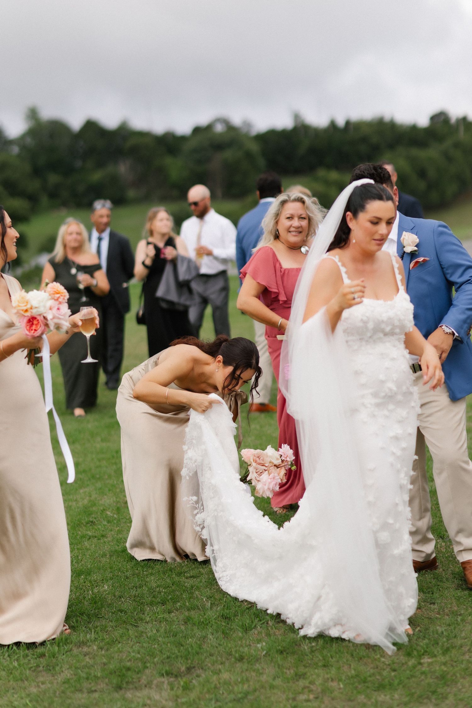 Bridesmaid adjusting bride's white lace wedding dress train on green grass at outdoor wedding reception.