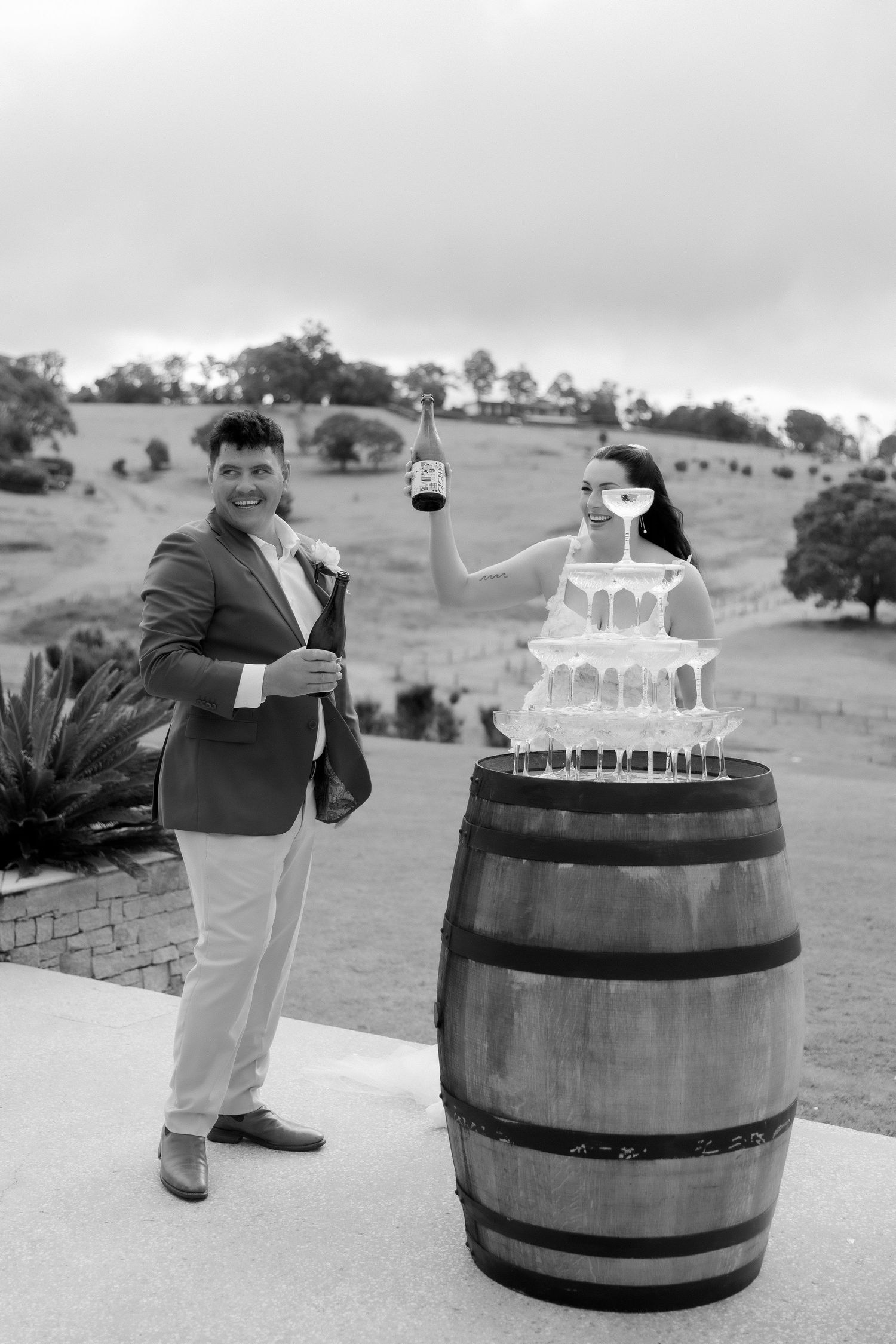 Black and white photo of couple pouring champagne tower on rustic wooden barrel at wedding reception.
