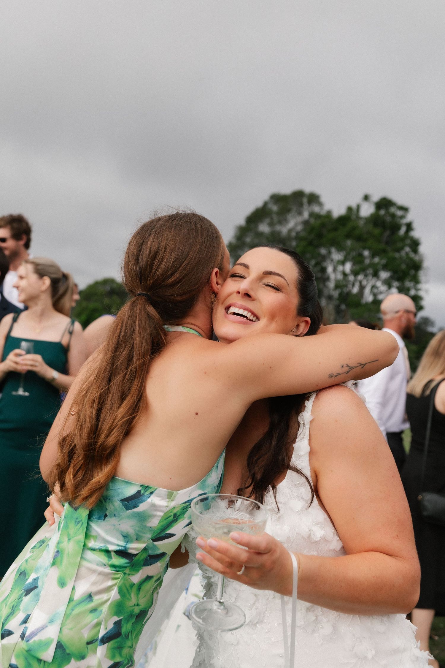 Joyful bride in white lace gown embracing a guest in green floral dress at outdoor wedding.