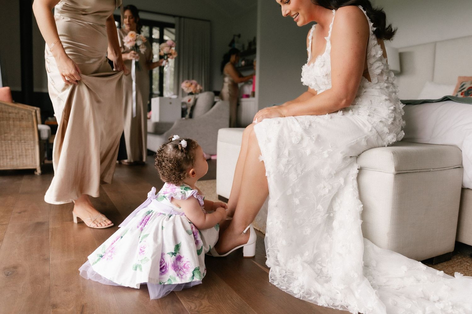 A flower girl helps the bride put on her wedding shoes while bridesmaids stand nearby in a bright room.