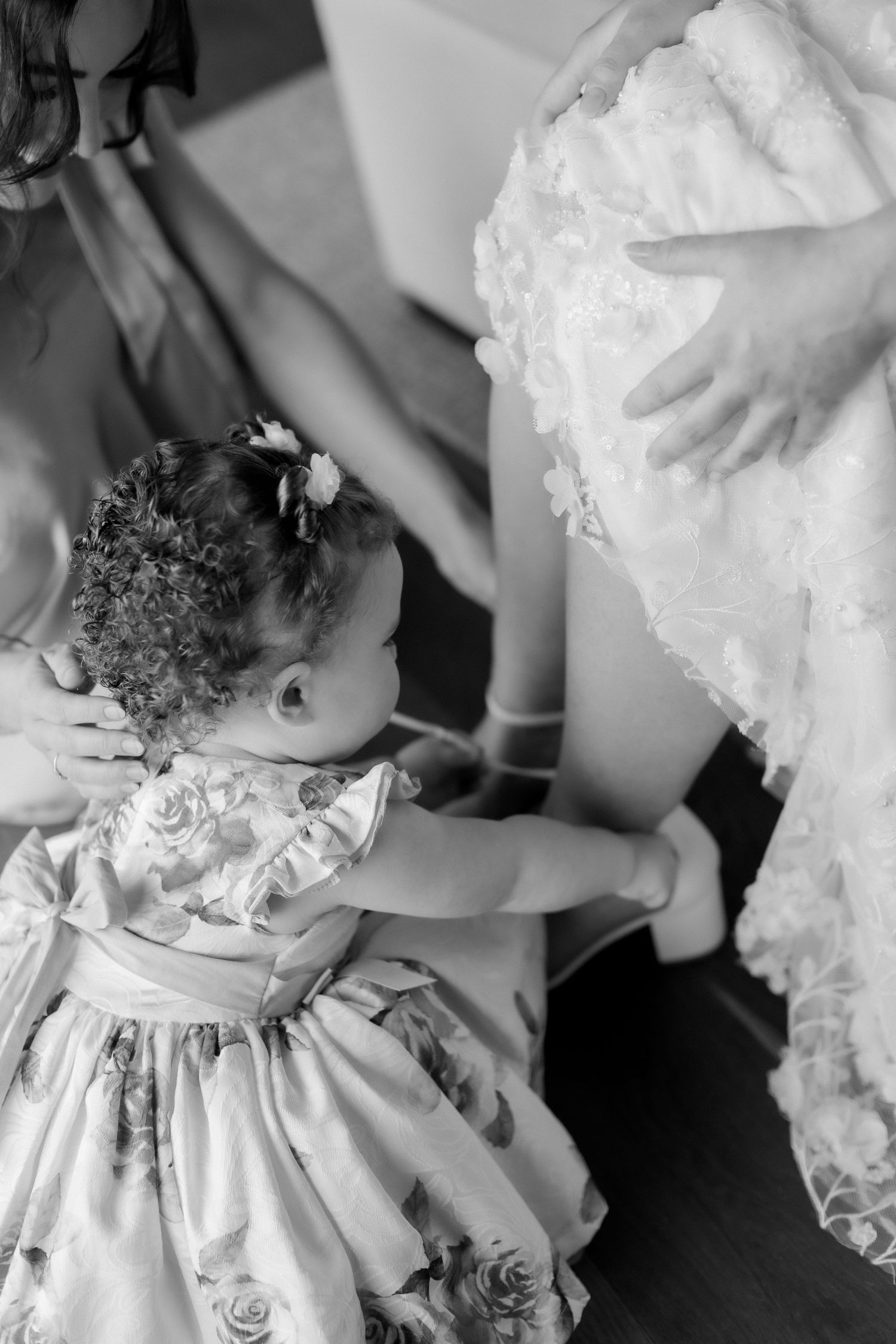 Black and white photo of a young flower girl in floral dress touching a brides lace wedding gown.