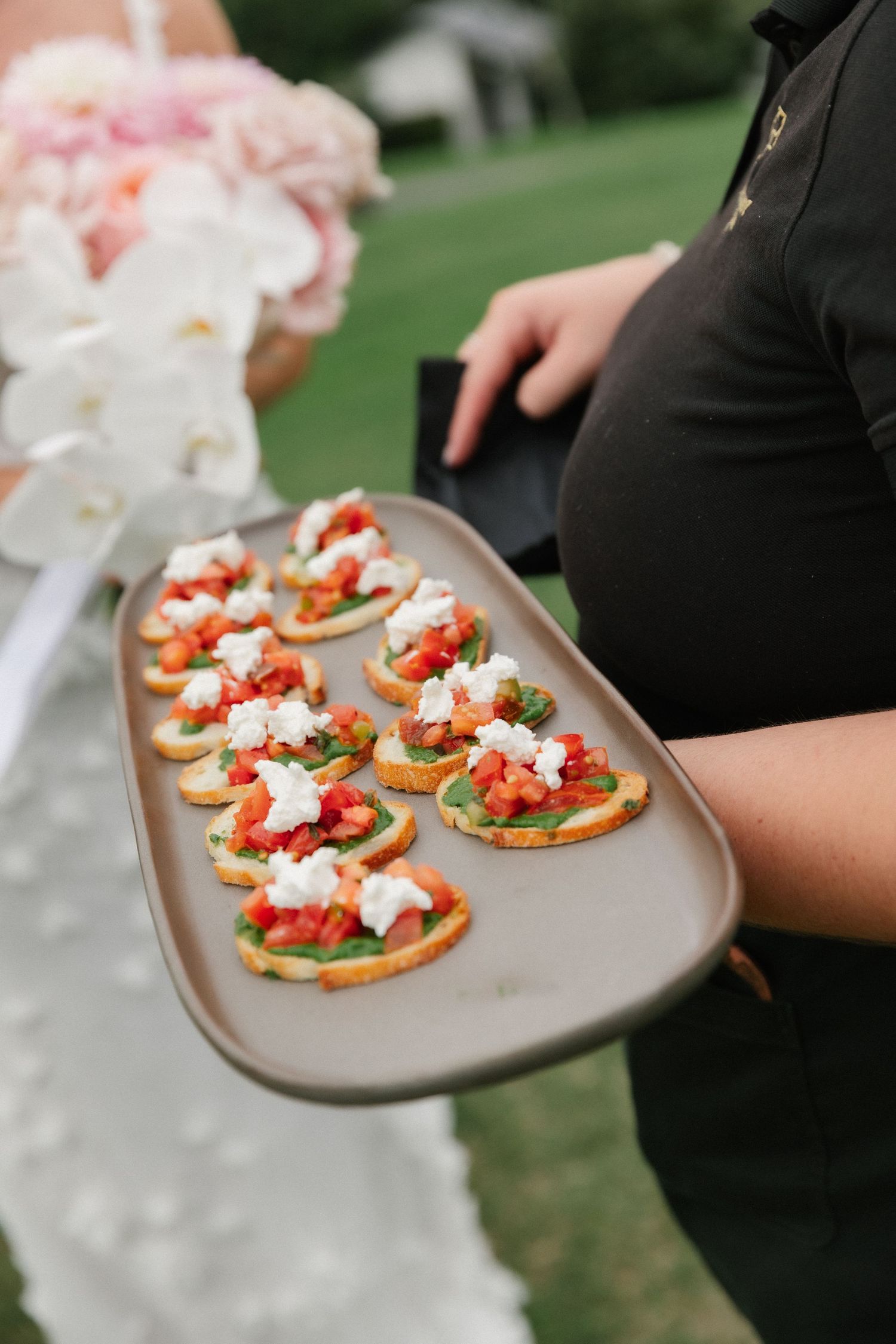 Server presenting tray of bruschetta topped with tomatoes and ricotta at outdoor wedding reception.